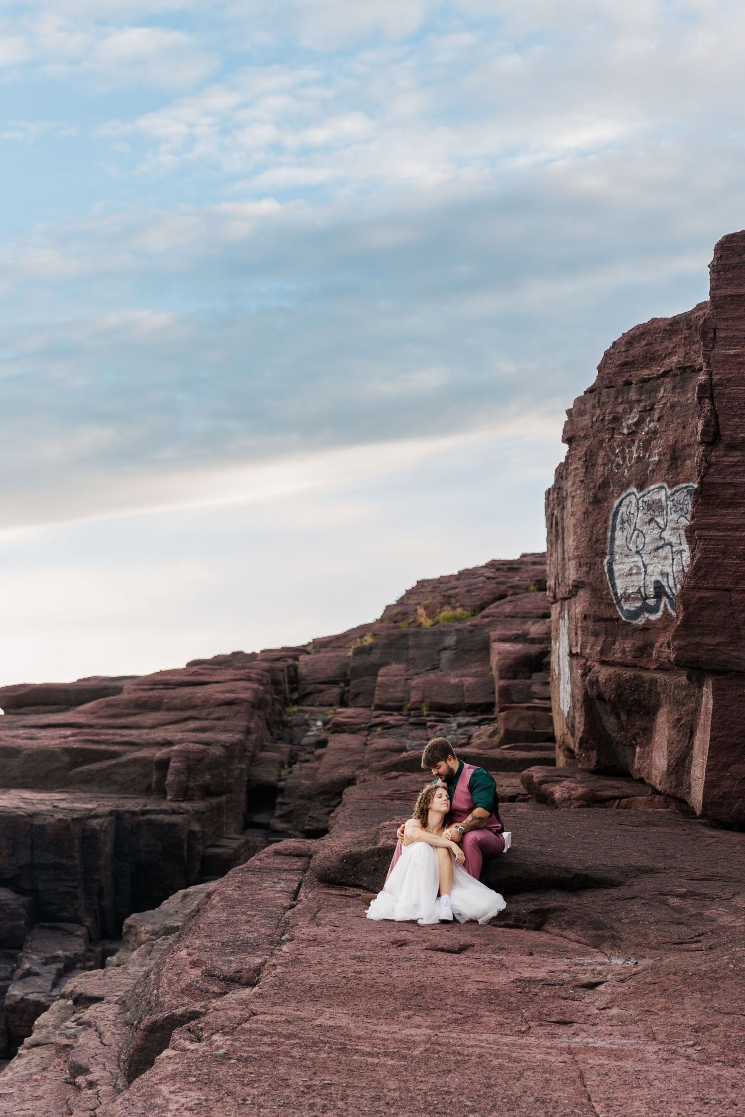 Two people share an intimate moment on red rock cliffs at sunset against a cloudy desert sky.