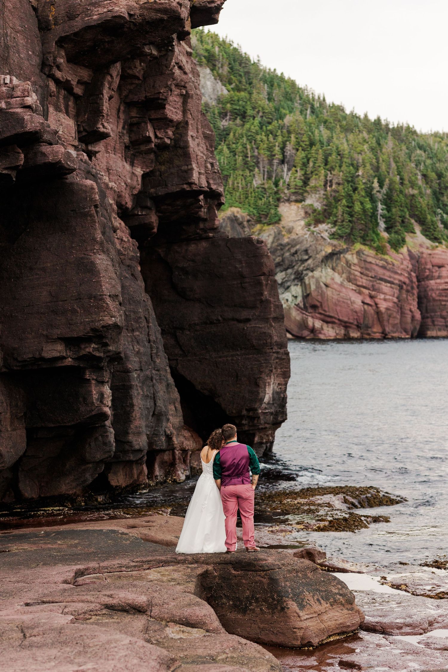 A pair stands near rugged red cliffs overlooking the ocean at sunset along a rocky coastline.