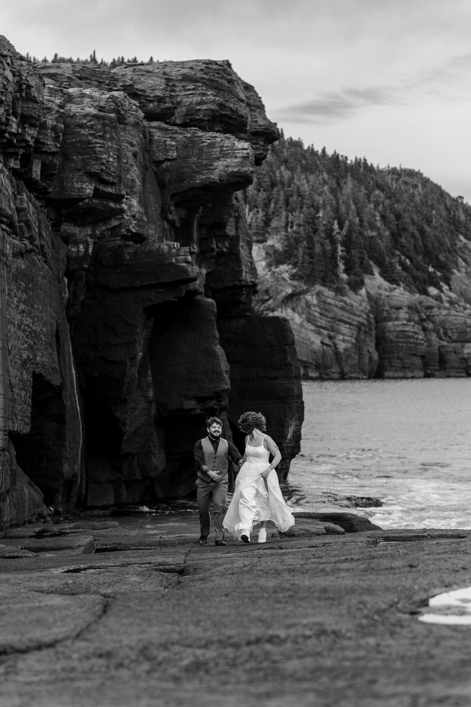 Black and white photo of a couple by rugged coastal cliffs with crashing waves and forest backdrop.