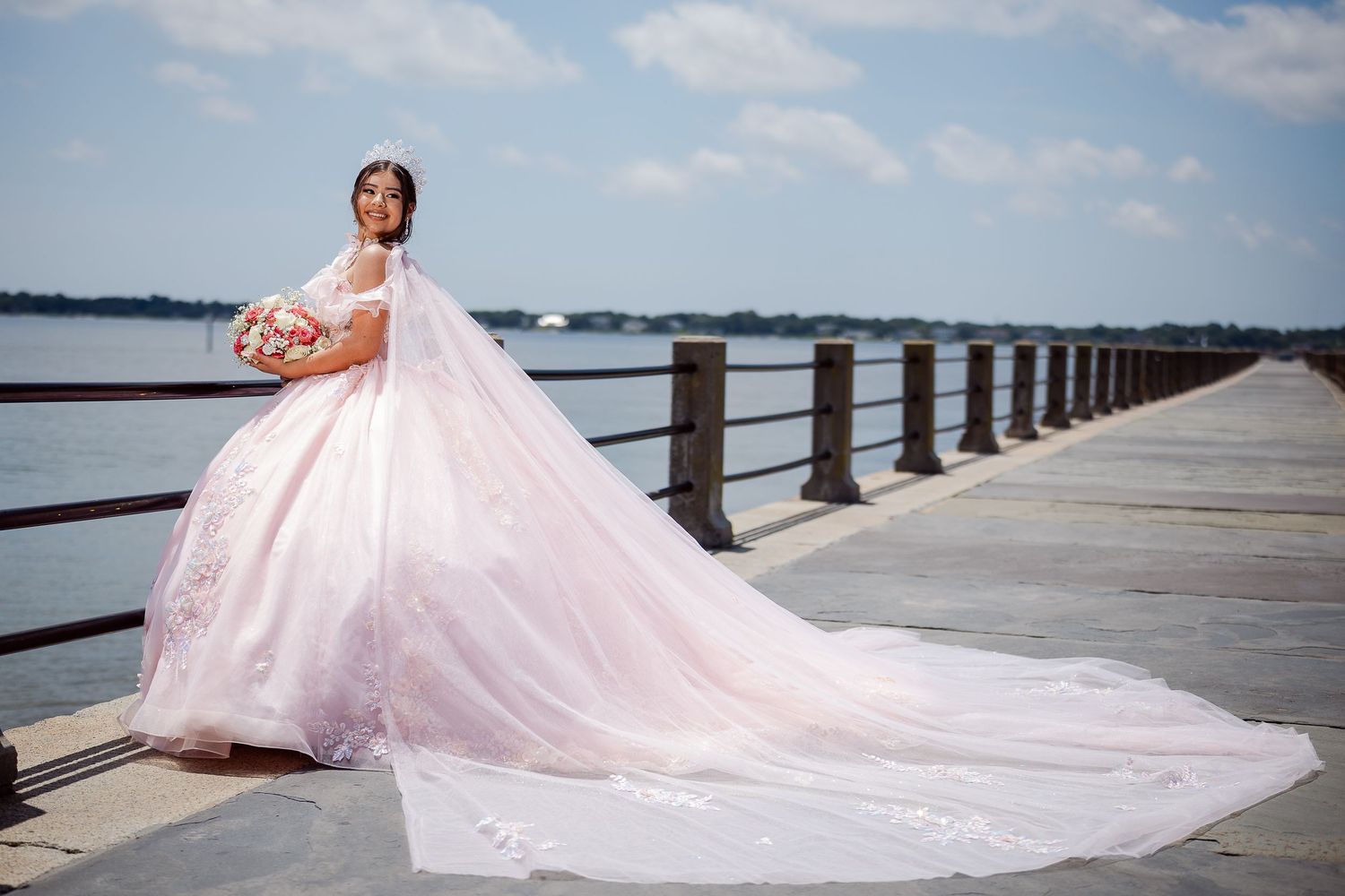 A bride in a pale pink ball gown with lace details poses beside ornate white stone columns on a sunny day.