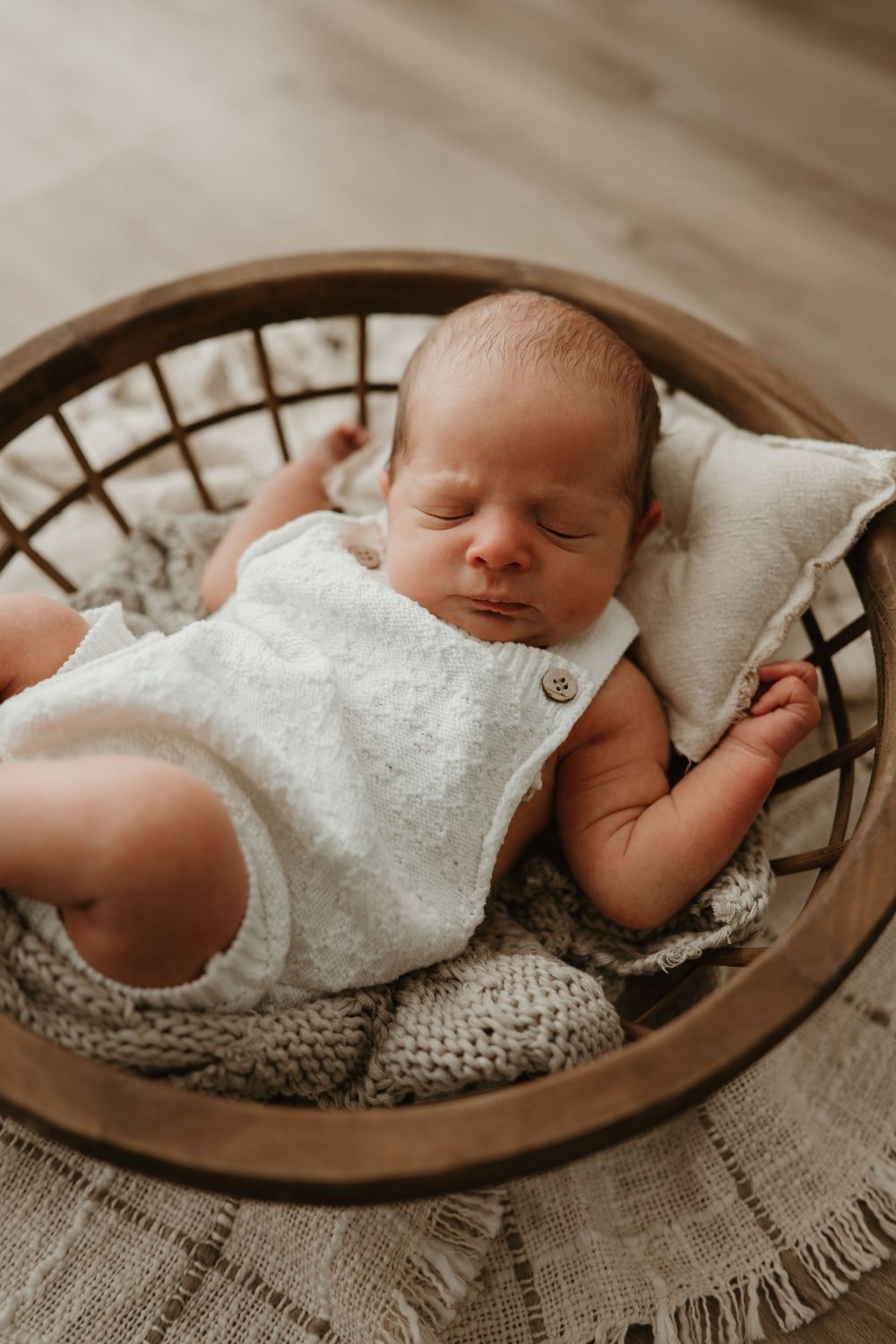 Sleeping newborn baby in white knit romper resting peacefully in a round wooden basket with soft blankets.
