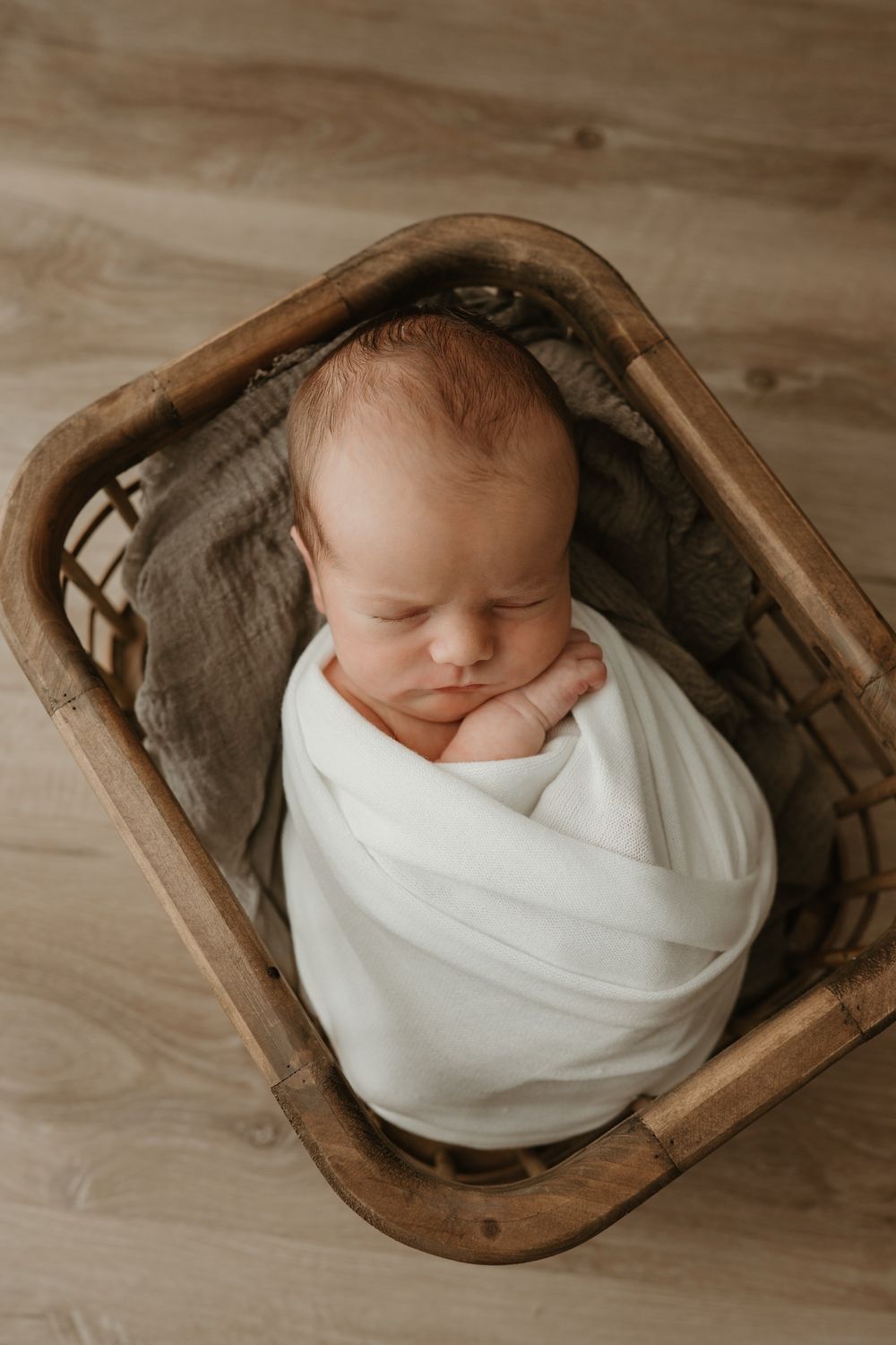 Newborn baby swaddled in white wrap sleeping peacefully in a rustic wooden basket with gray linen. Southern Saskatchewan newborn studio