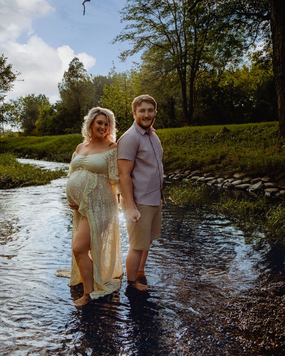 Couple standing together in shallow river water surrounded by trees and natural scenery at sunset.