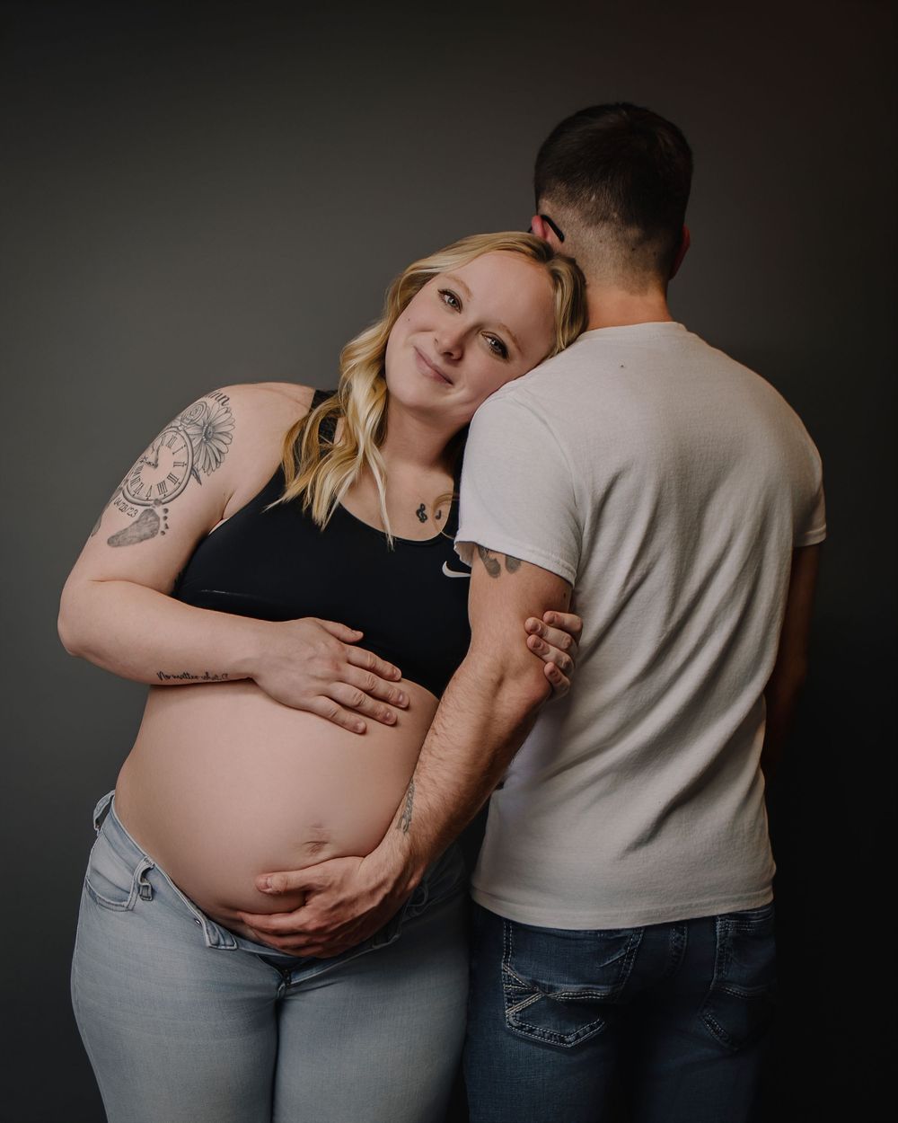 Intimate maternity portrait showing an expecting mother in casual attire embracing another person against a dark backdrop.