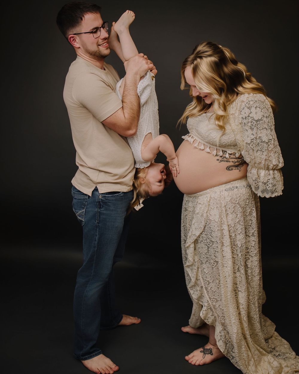 A couple poses for a pregnancy photo, with one holding a toddler upside down while wearing a lace maternity gown.