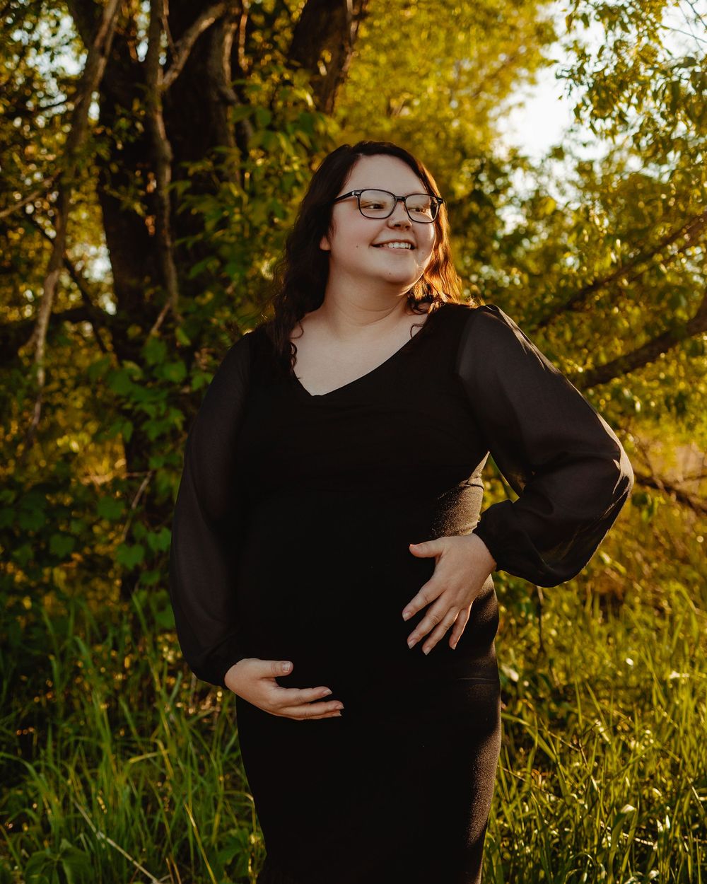 A person in a black dress stands outdoors during golden hour with autumn foliage in the background.