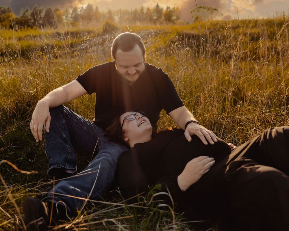 Blissful couple shares a romantic moment lying in golden sunlit grass at sunset during an engagement photoshoot.
