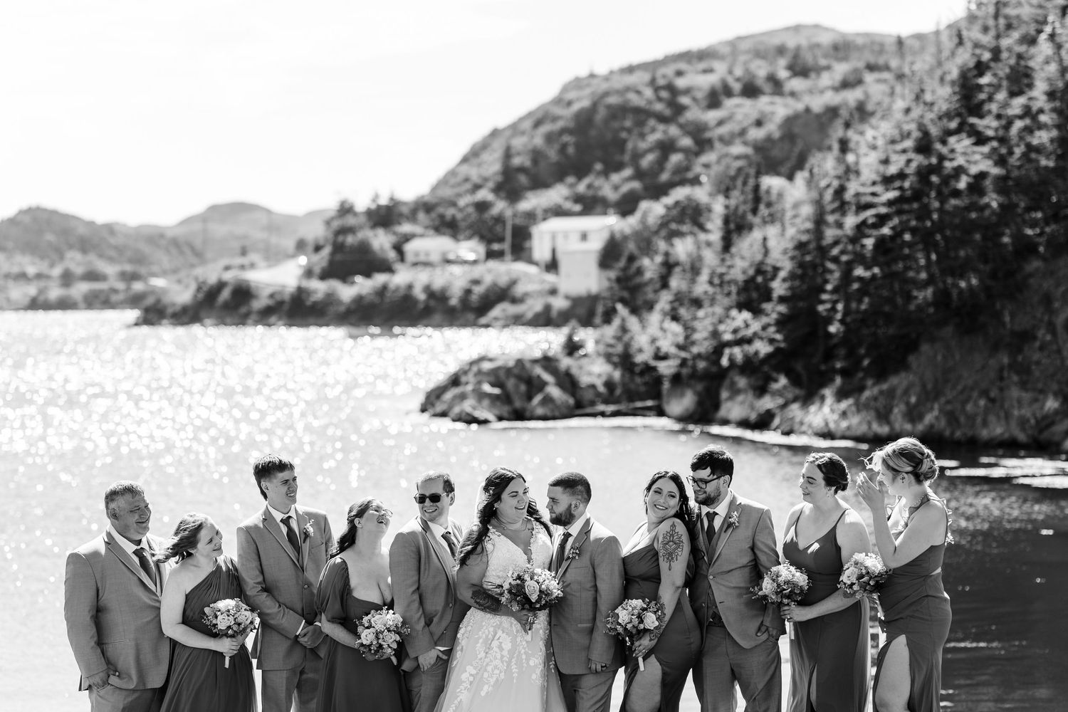 A group in formalwear stands together near a scenic waterfront landscape with cliffs and mountains in the background.