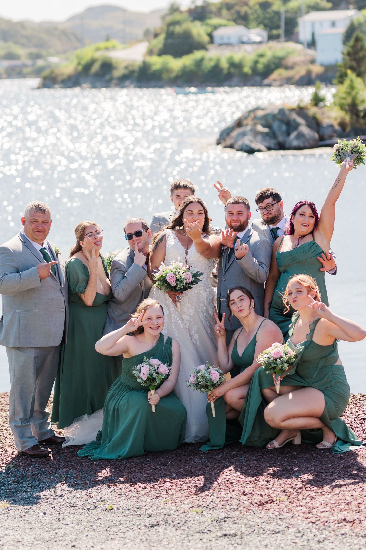 A wedding party in gray and emerald green attire poses casually together on a rocky beach with water in the background.
