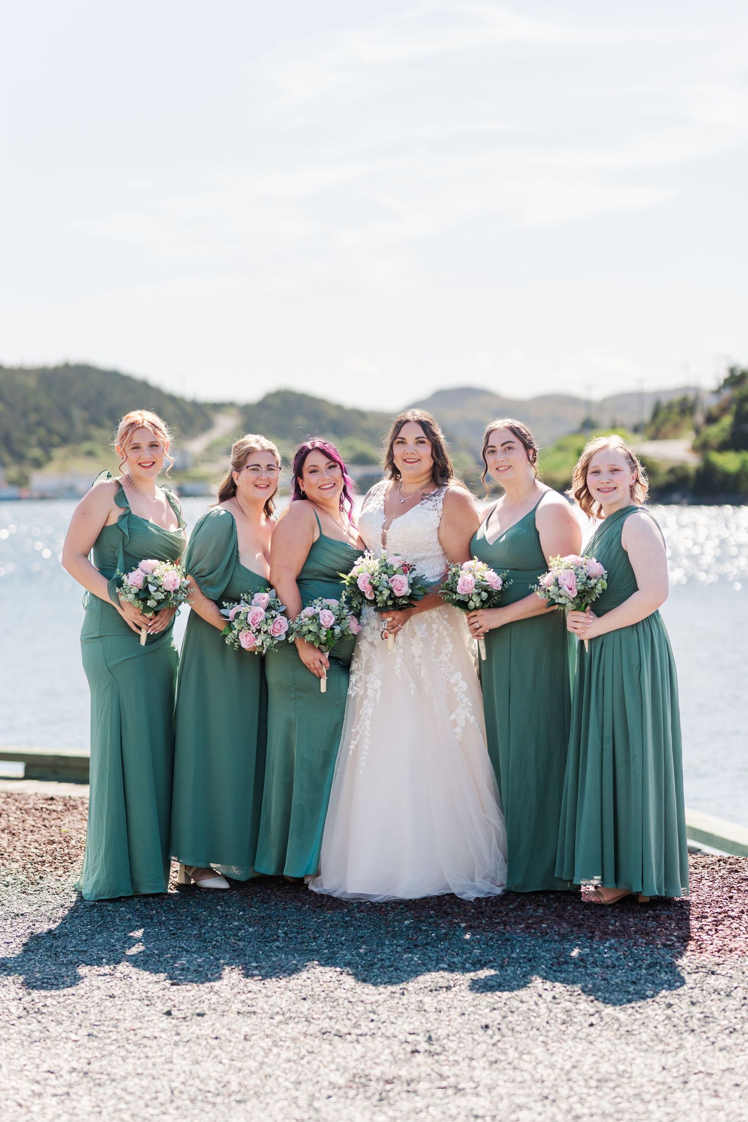 A bridal party in emerald green dresses and white wedding gown poses together on a rocky beach shoreline by the water.