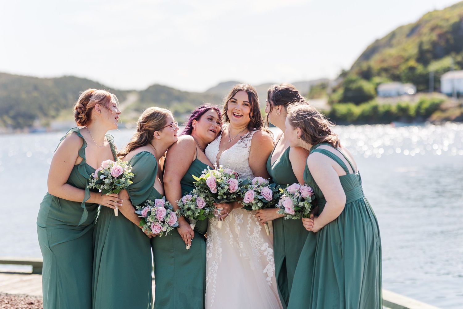 Bridesmaids in matching sage green dresses embrace and laugh together on a sunny waterfront.