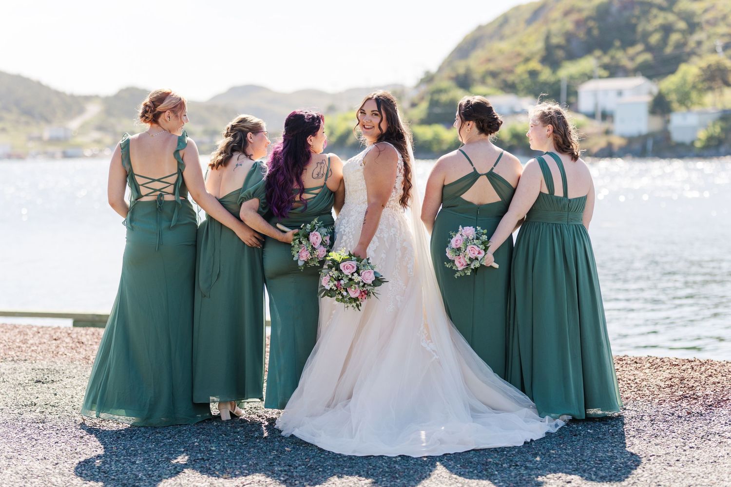 Bridal party in matching emerald green bridesmaid dresses stand with bride in white gown by waterfront on rocky beach.