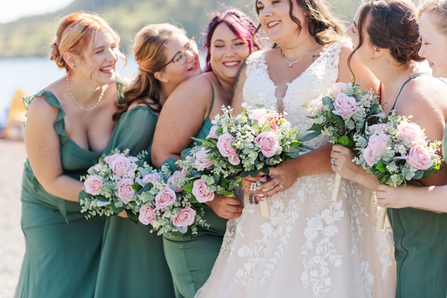 A group wearing elegant green bridesmaid dresses and white wedding gown hold pink and white floral bouquets in sunny outdoor setting.