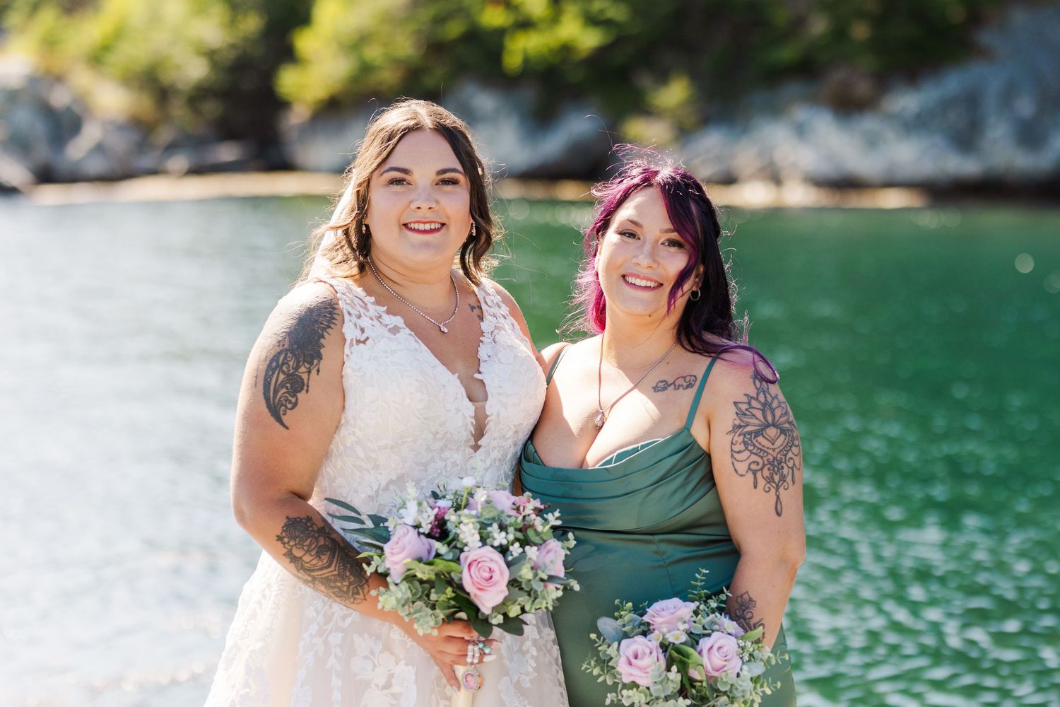 Two people holding purple and white floral bouquets pose together by a green lake on a sunny day.
