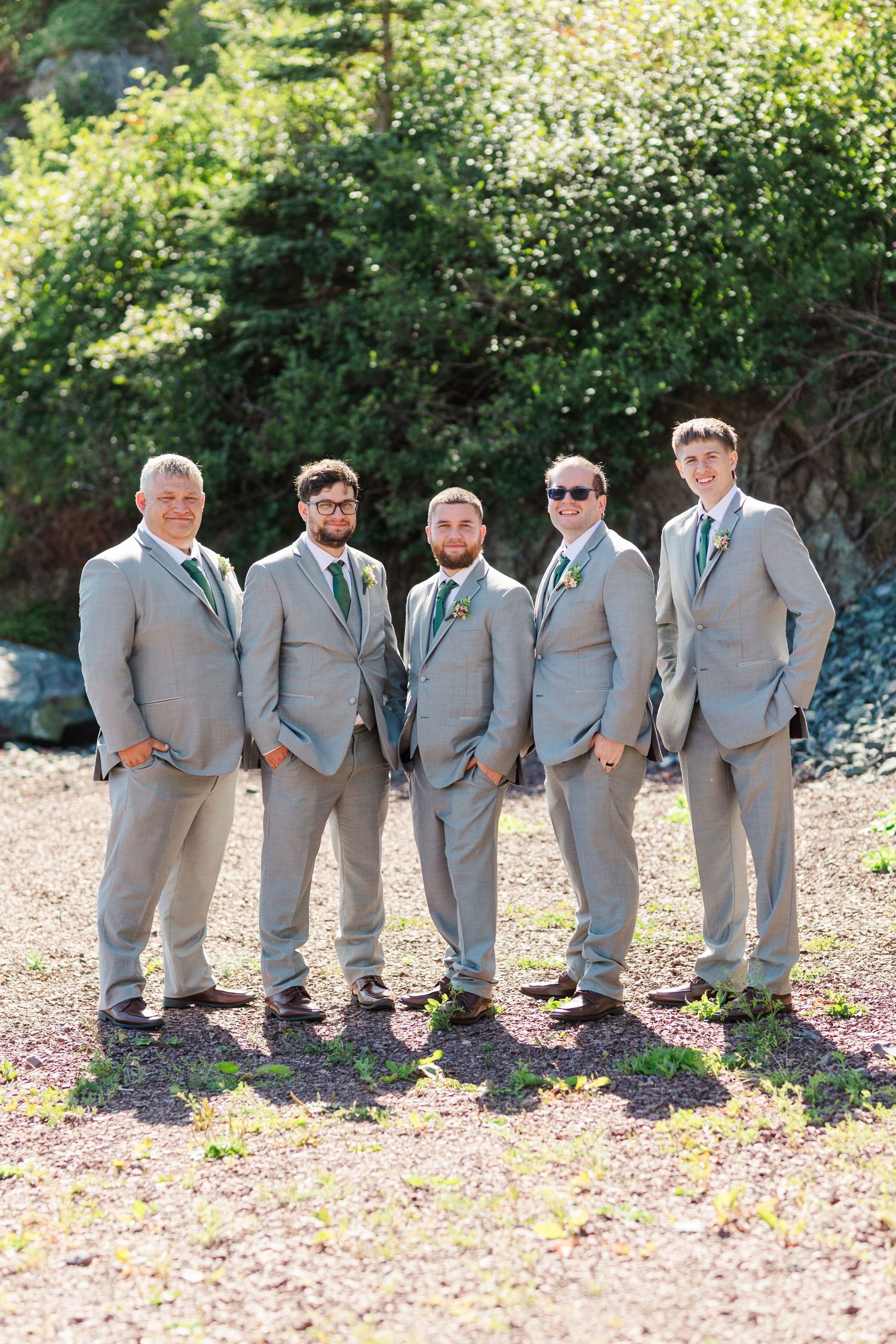 A sequence of groomsmen in matching gray suits perform a playful line dance pose on flower-covered ground.