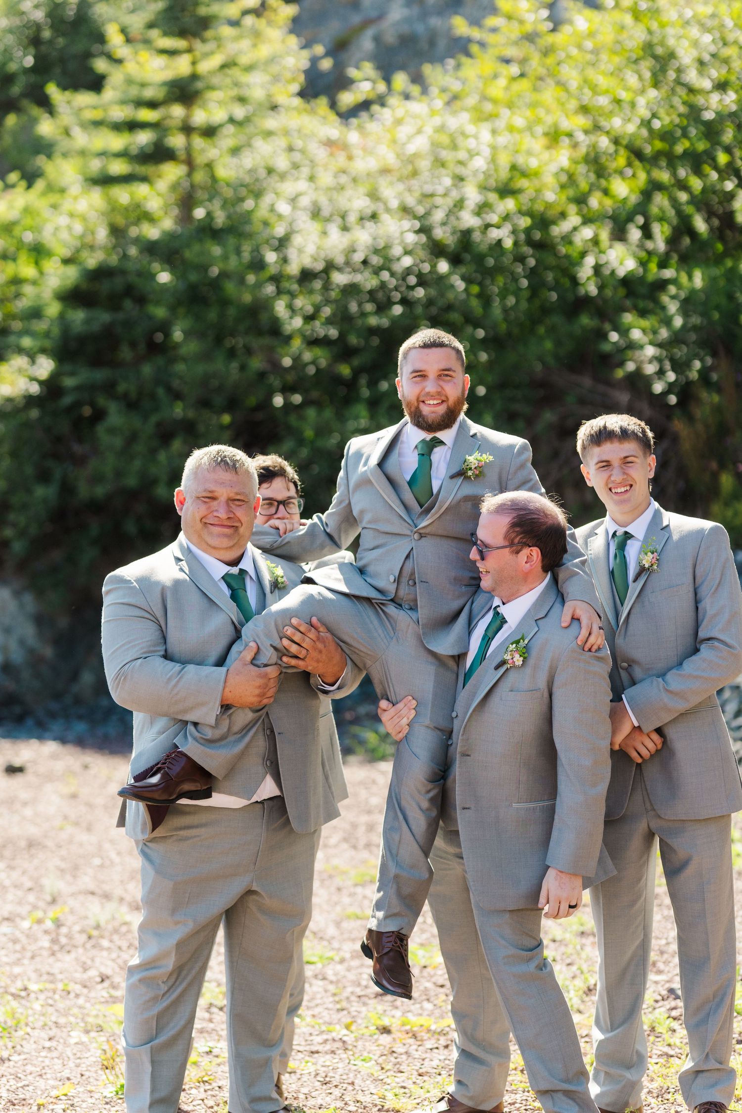 Groomsmen in gray suits posing playfully and candidly during wedding photos outdoors with trees in the background.