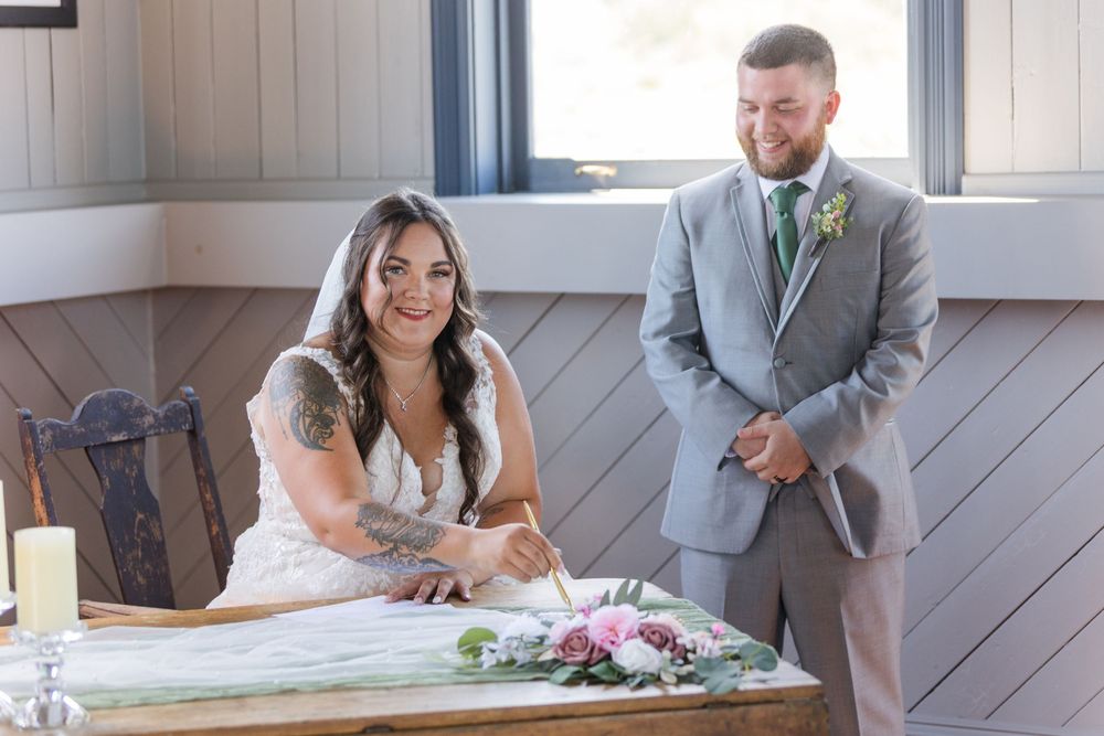 People signing documents at a wedding ceremony in a rustic barn venue with gray wood paneling.