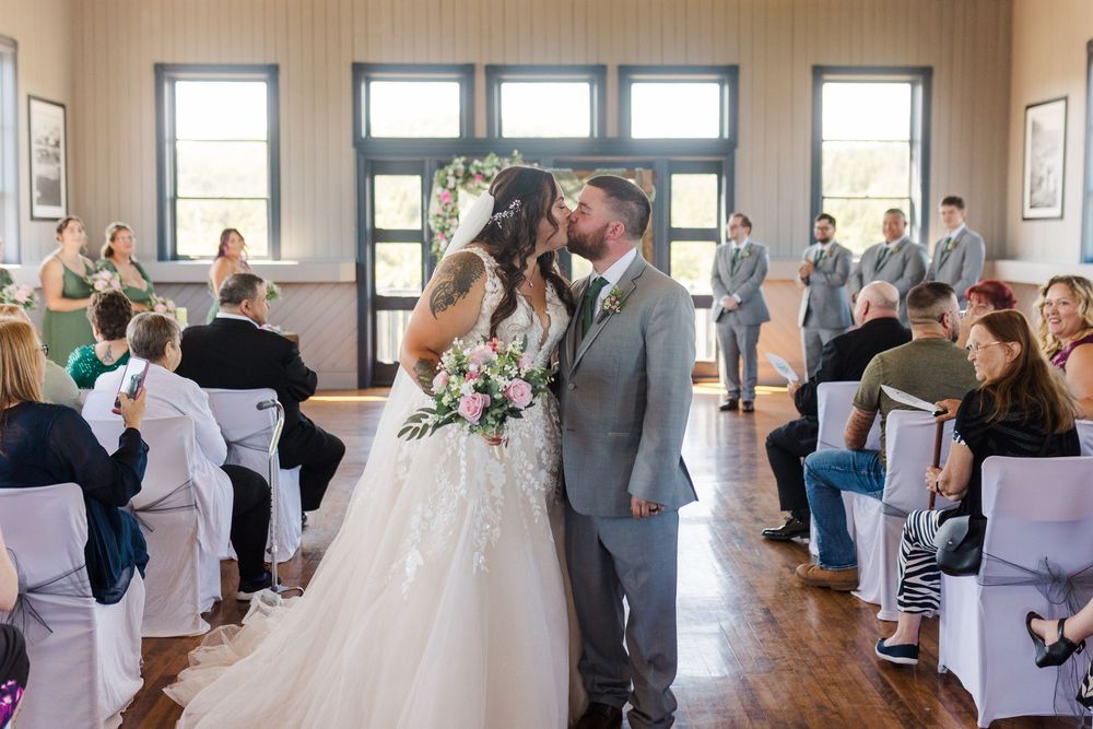 A couple shares their first kiss during an indoor wedding ceremony surrounded by seated guests in a bright room.