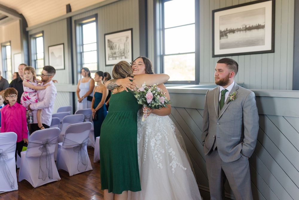Guests in formal attire greet and embrace at an intimate indoor wedding ceremony with natural lighting and gray walls.