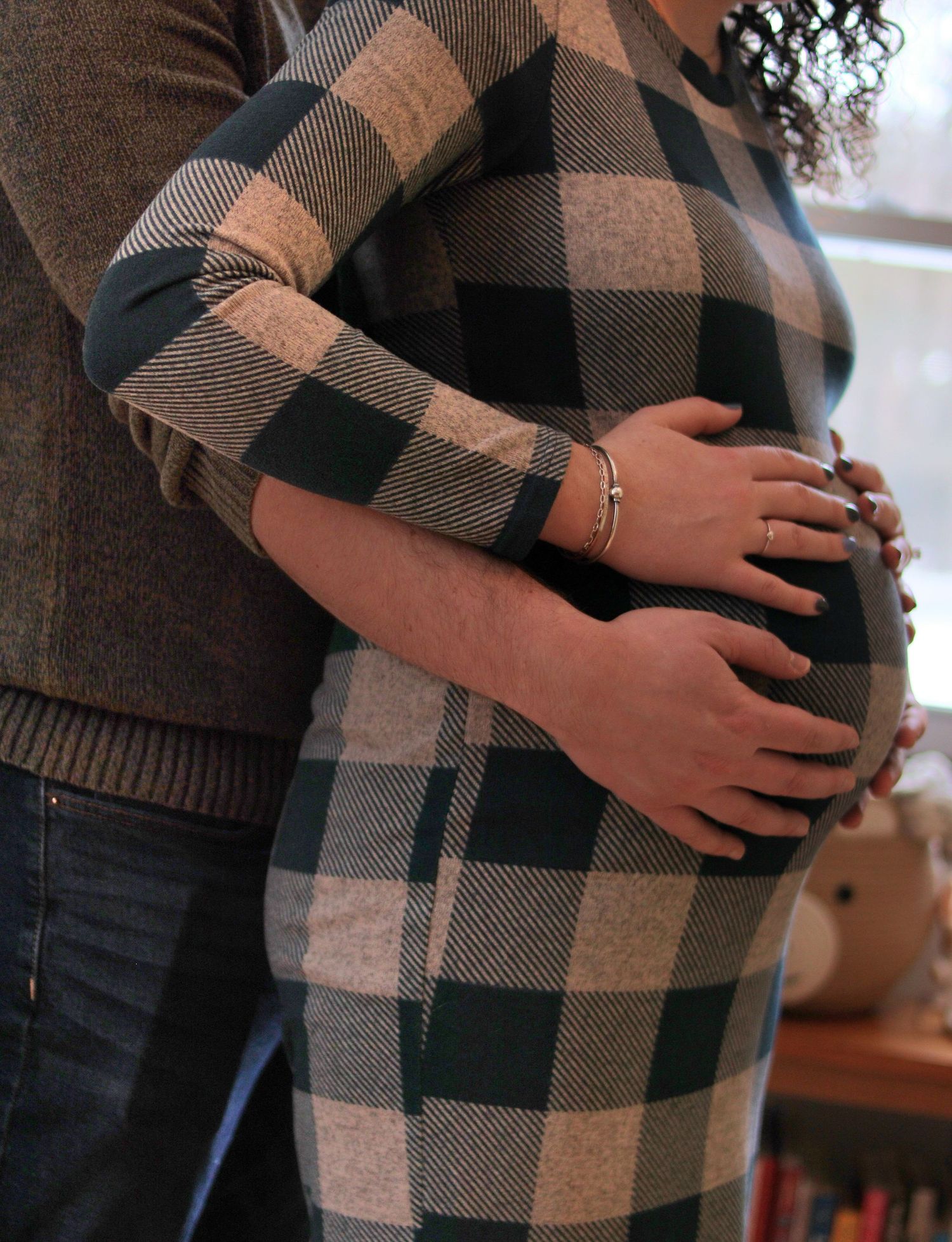 A pregnant couple embraces indoors, both cradling the baby bump in a plaid maternity dress.