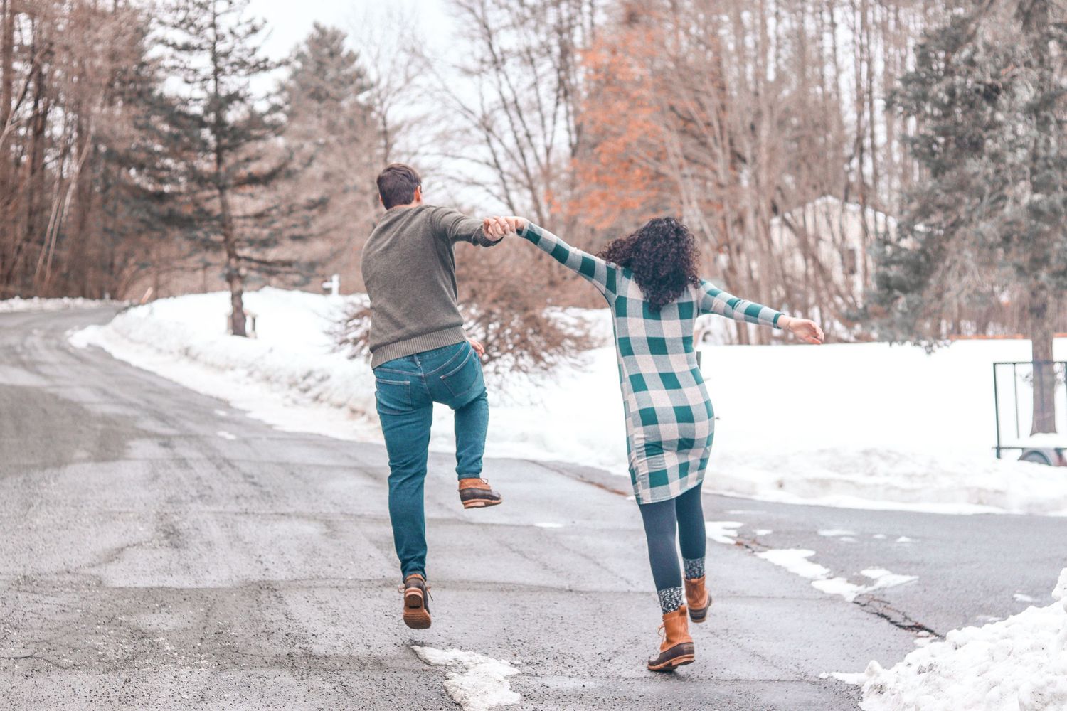A couple holds hands and dances playfully on a snowy road surrounded by winter trees.