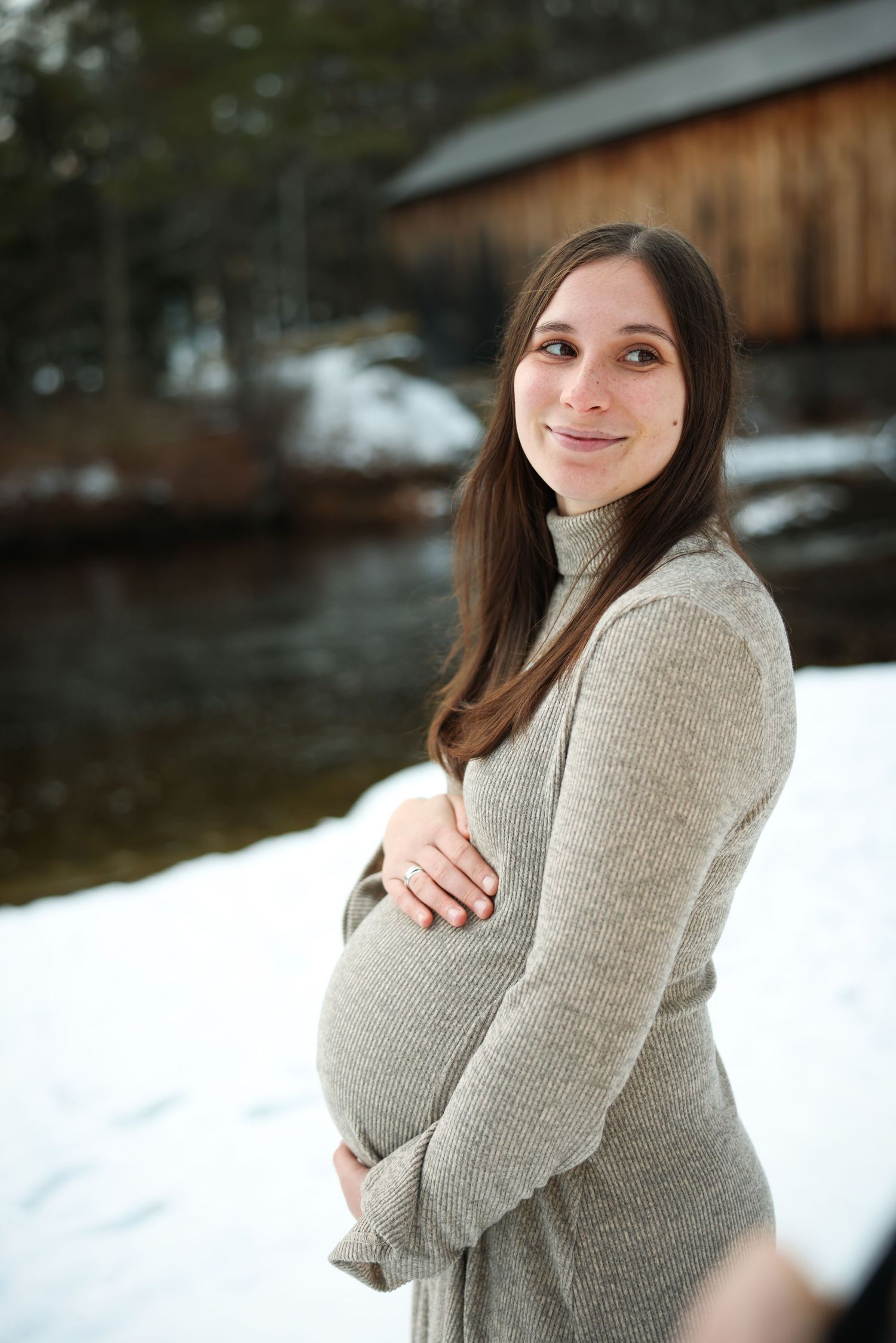 Pregnant woman in gray dress cradling baby bump by snowy riverside in winter.