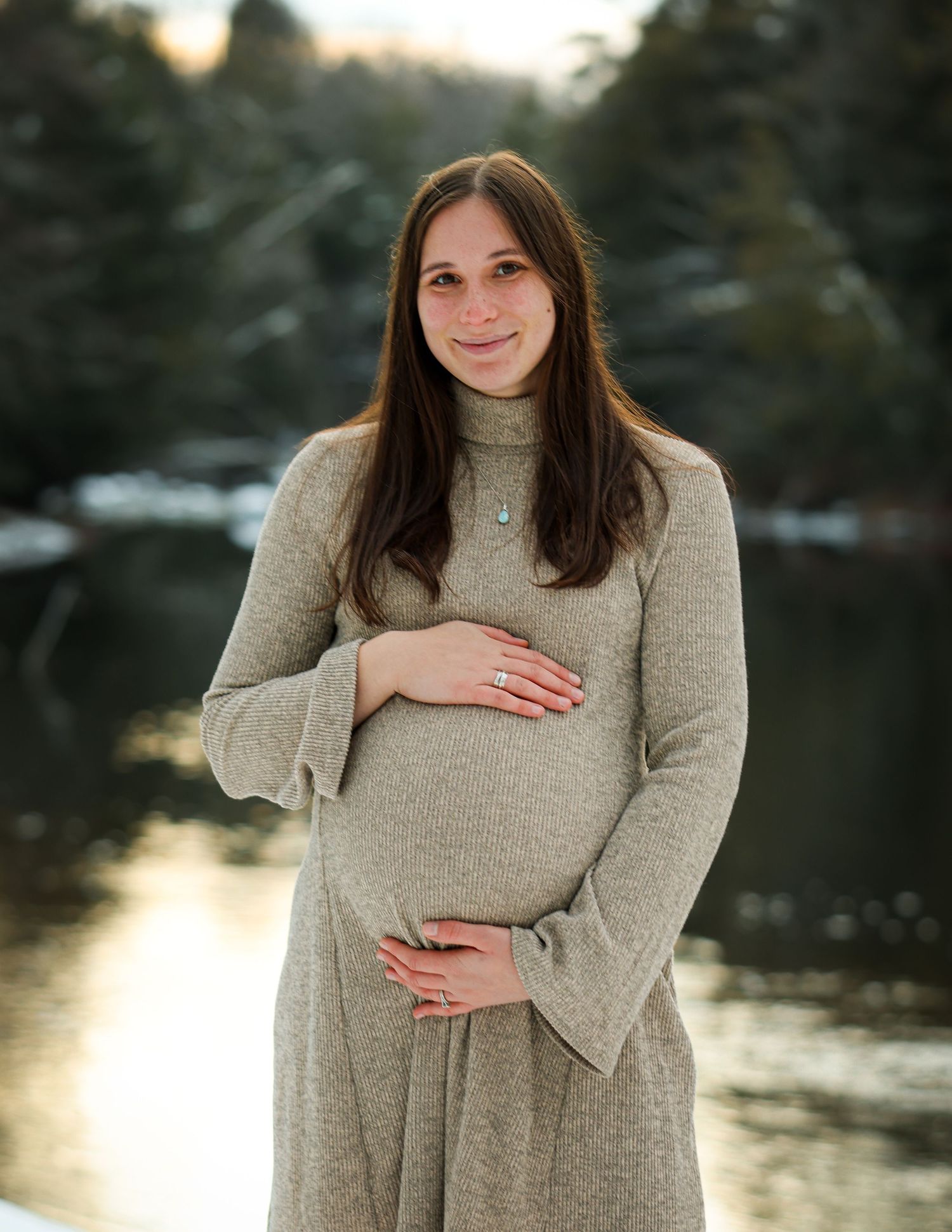 Smiling pregnant woman in beige knit dress holding baby bump by snowy winter river.