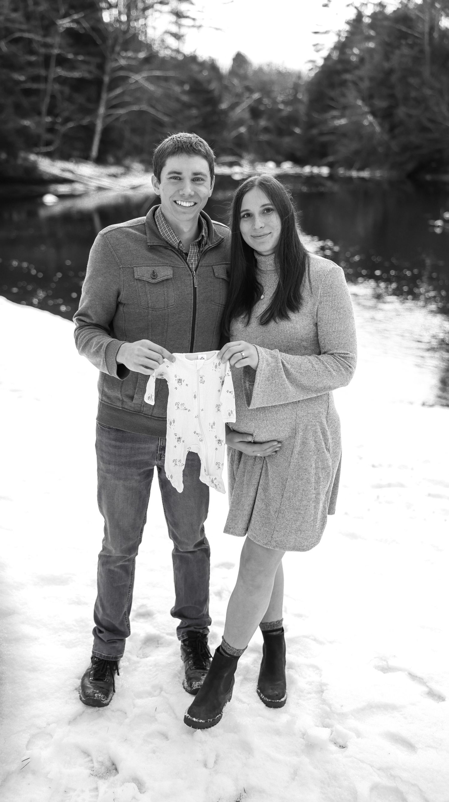 Couple holding baby onesie in snowy winter landscape announcing pregnancy in black and white.