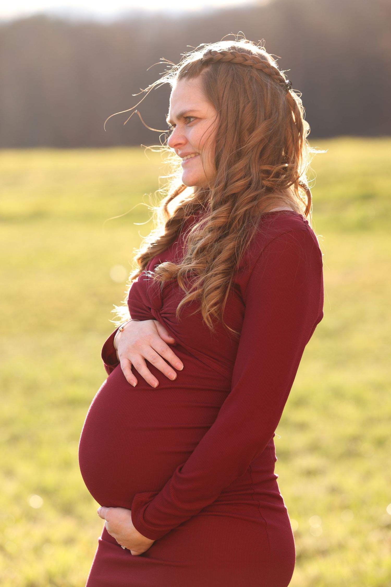 Pregnant woman in burgundy dress holding belly outdoors in golden sunlight with curly hair.