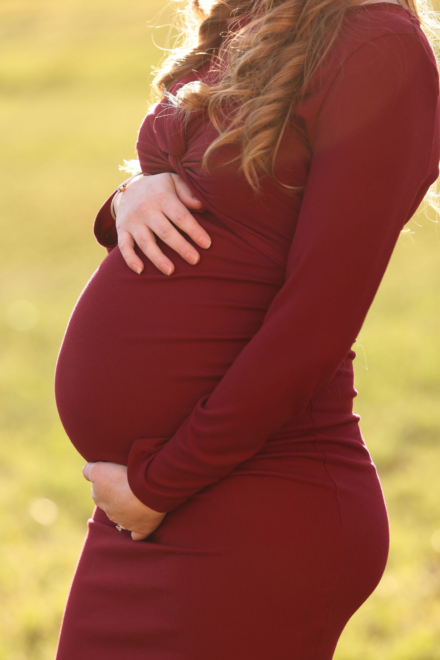 Close-up of pregnant woman in burgundy dress gently holding baby bump outdoors in warm sunlight.