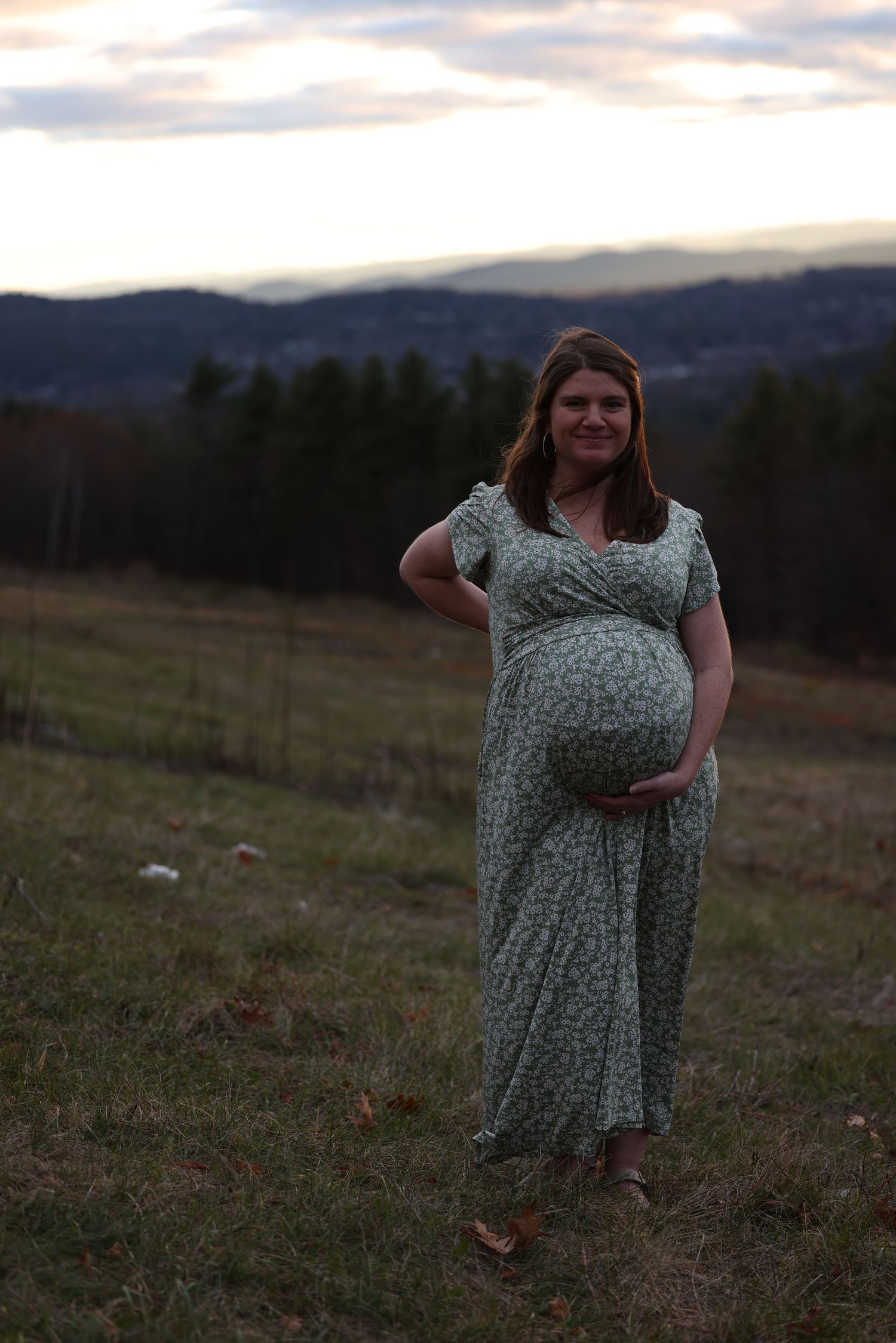 Smiling pregnant woman in floral dress standing in field at dusk with rolling hills behind her.
