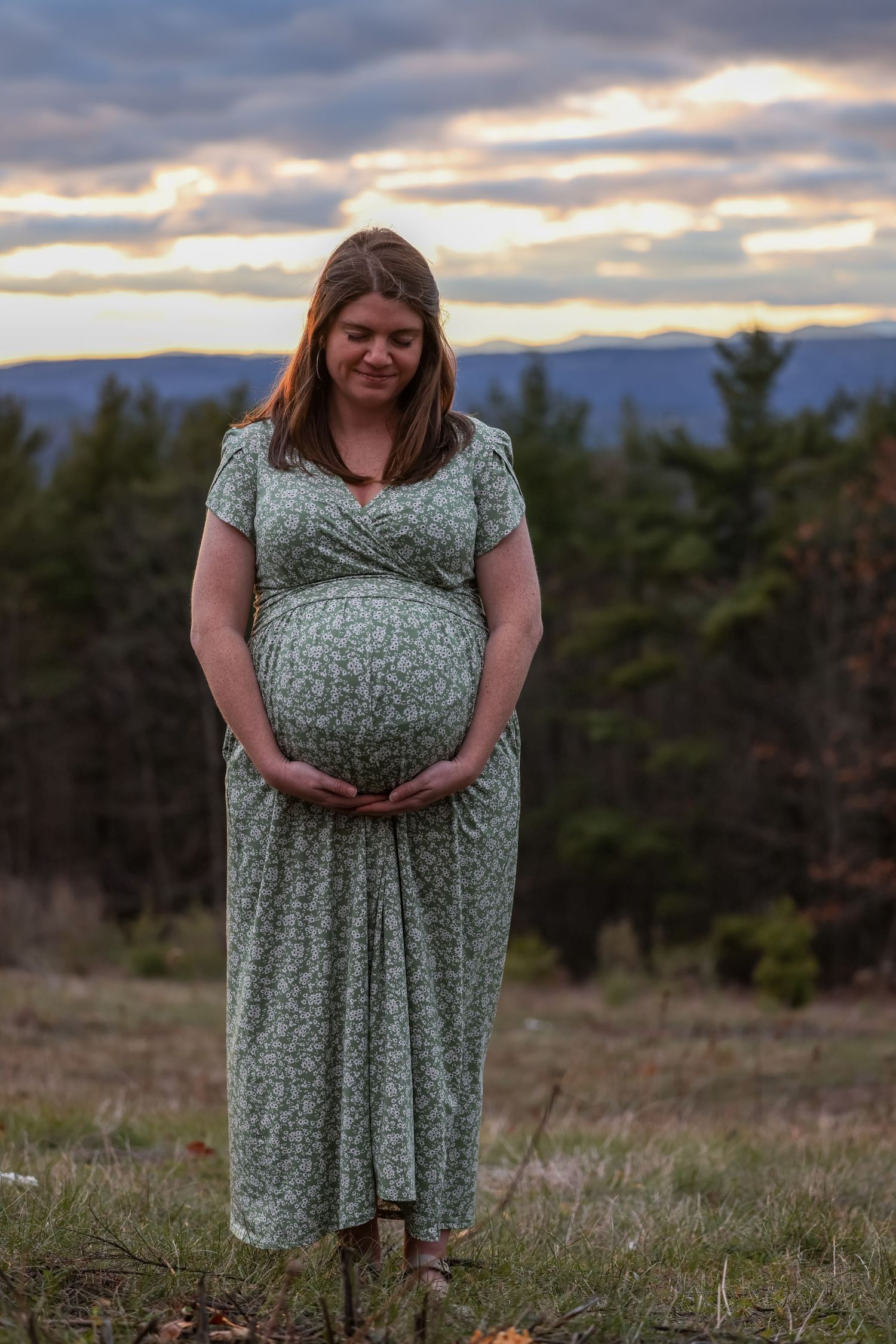 Pregnant woman in floral dress holding baby bump outdoors at sunset with mountain backdrop.