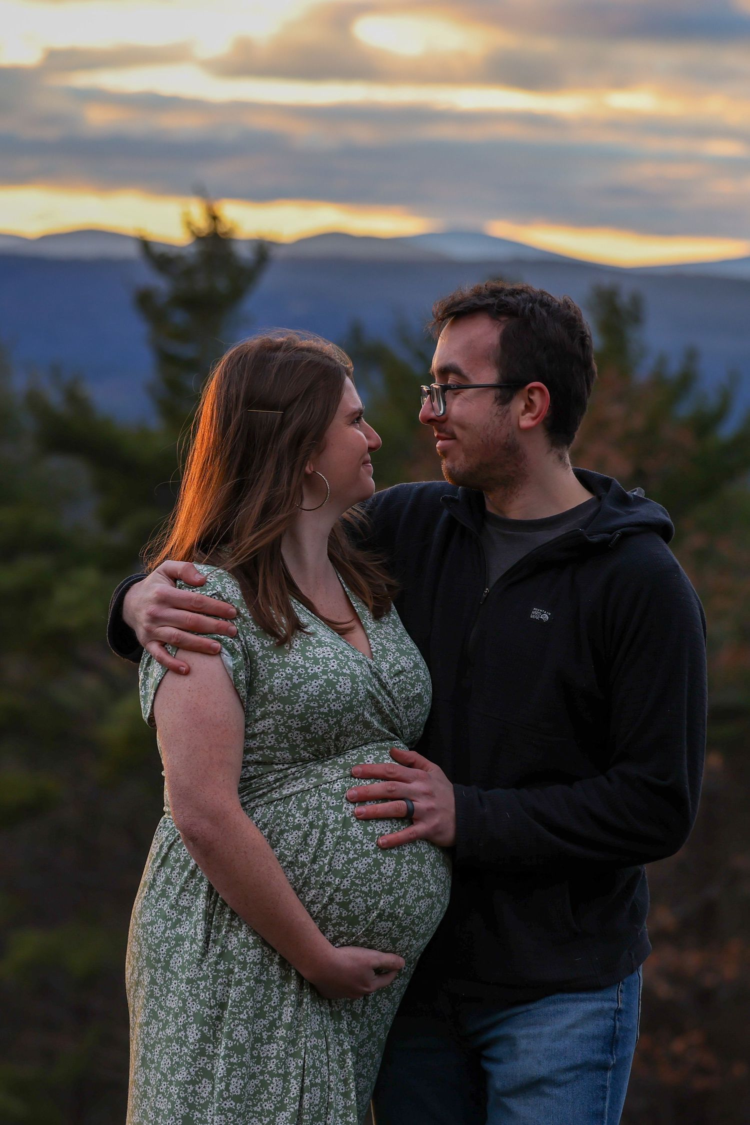 Couple sharing tender moment outdoors at sunset, man embracing pregnant woman in floral dress.