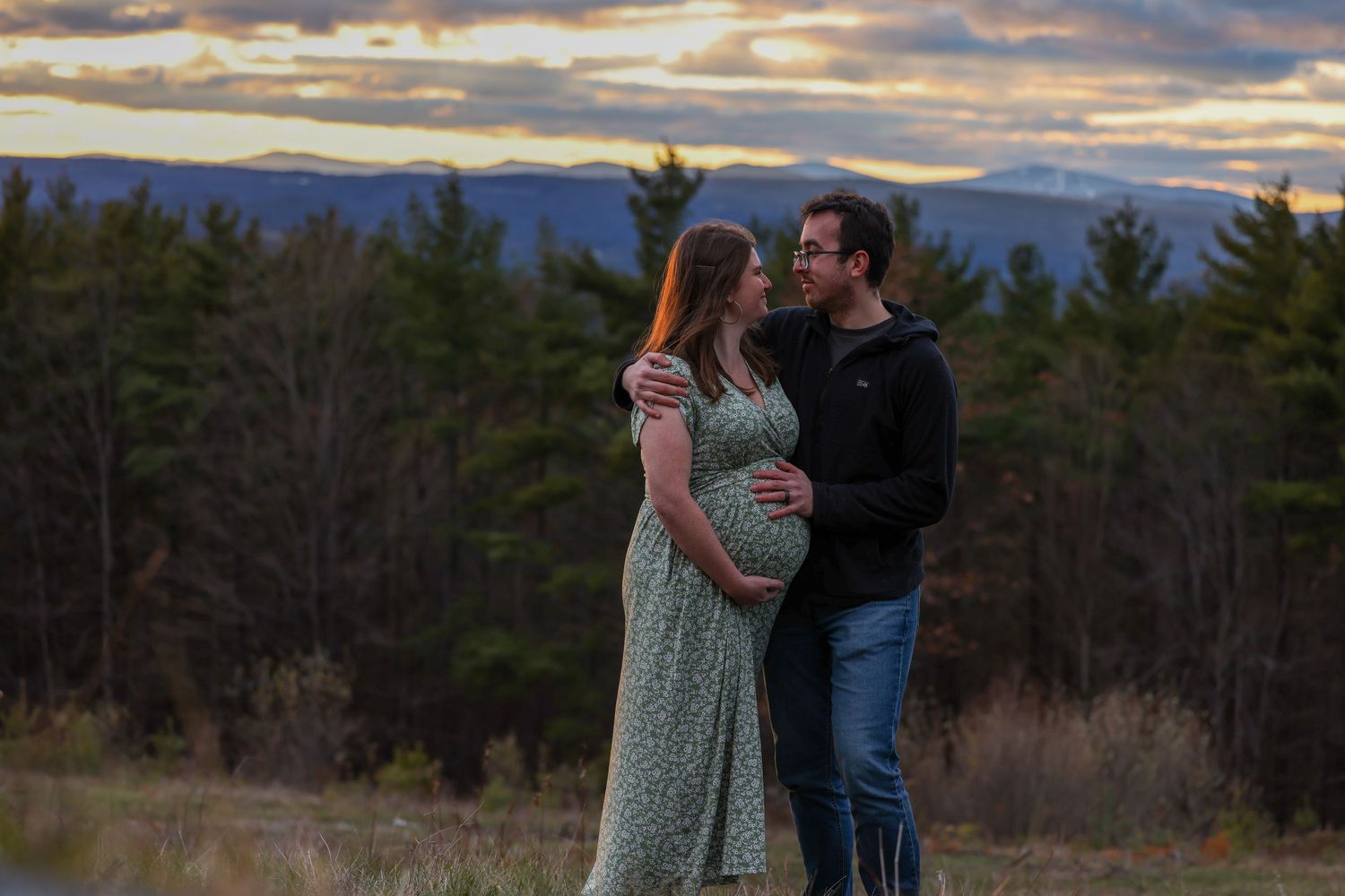 Couple standing together in field at dusk, man embracing pregnant woman with mountain views.
