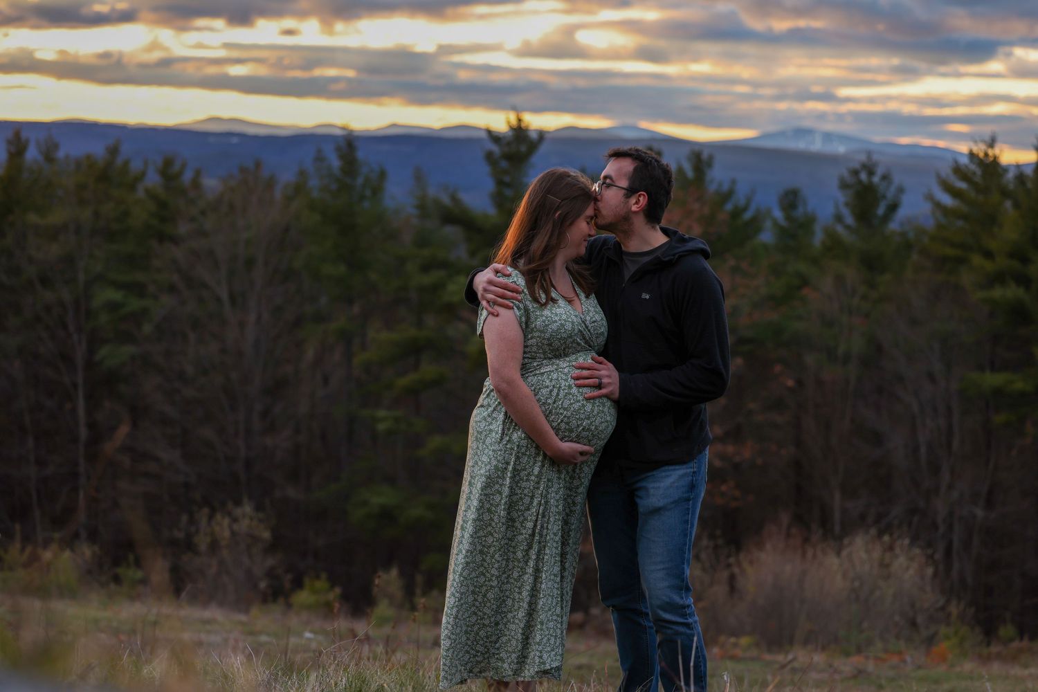 Man kissing pregnant woman's head at sunset in open field with mountain landscape behind them.