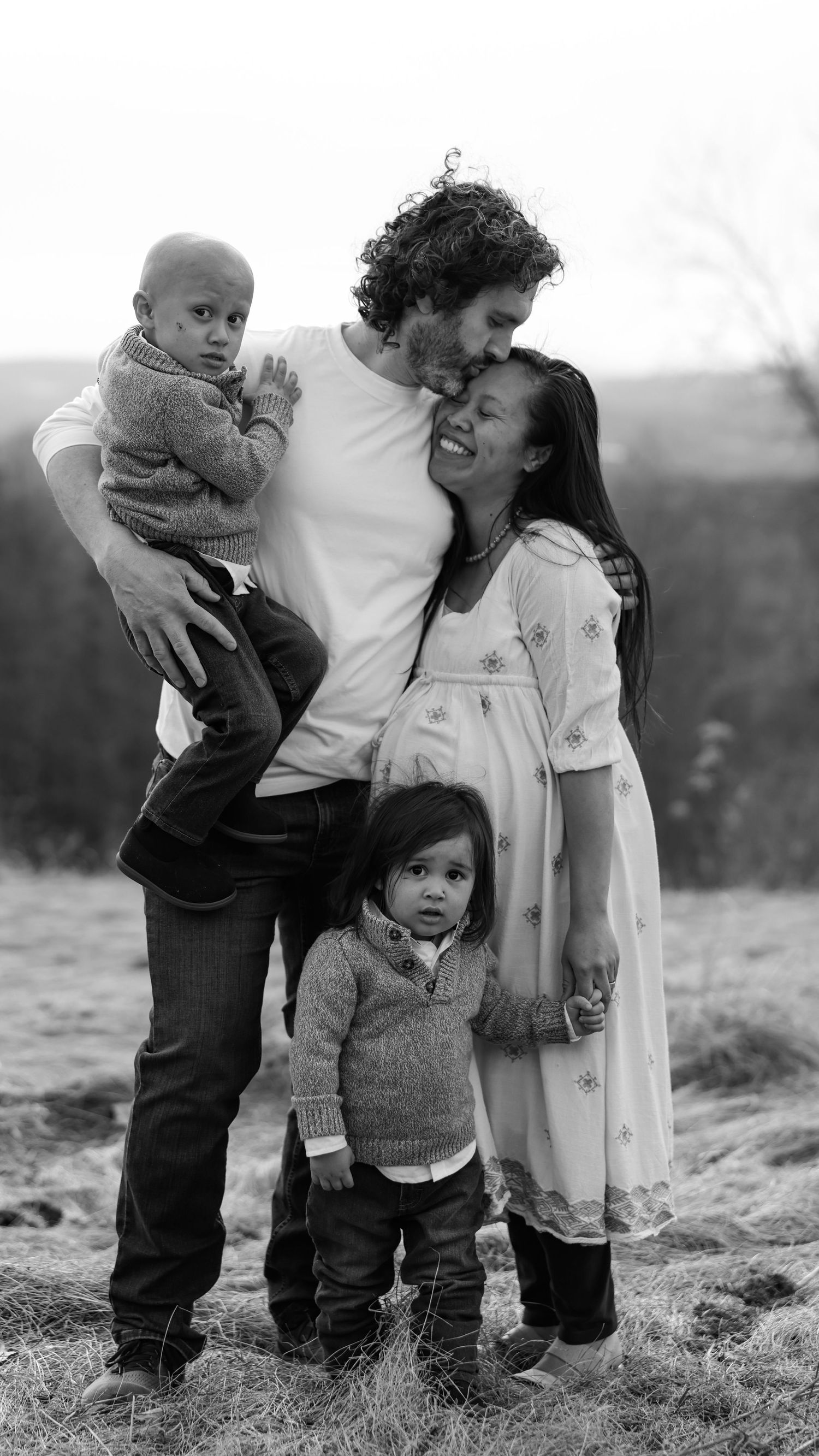 Black and white family portrait with father, pregnant mother, and two young children outdoors.