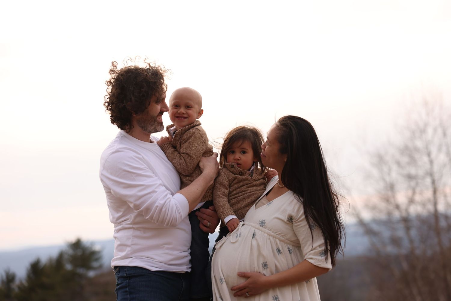 Happy family with pregnant mother, father, and two toddlers posing together at sunset outdoors.