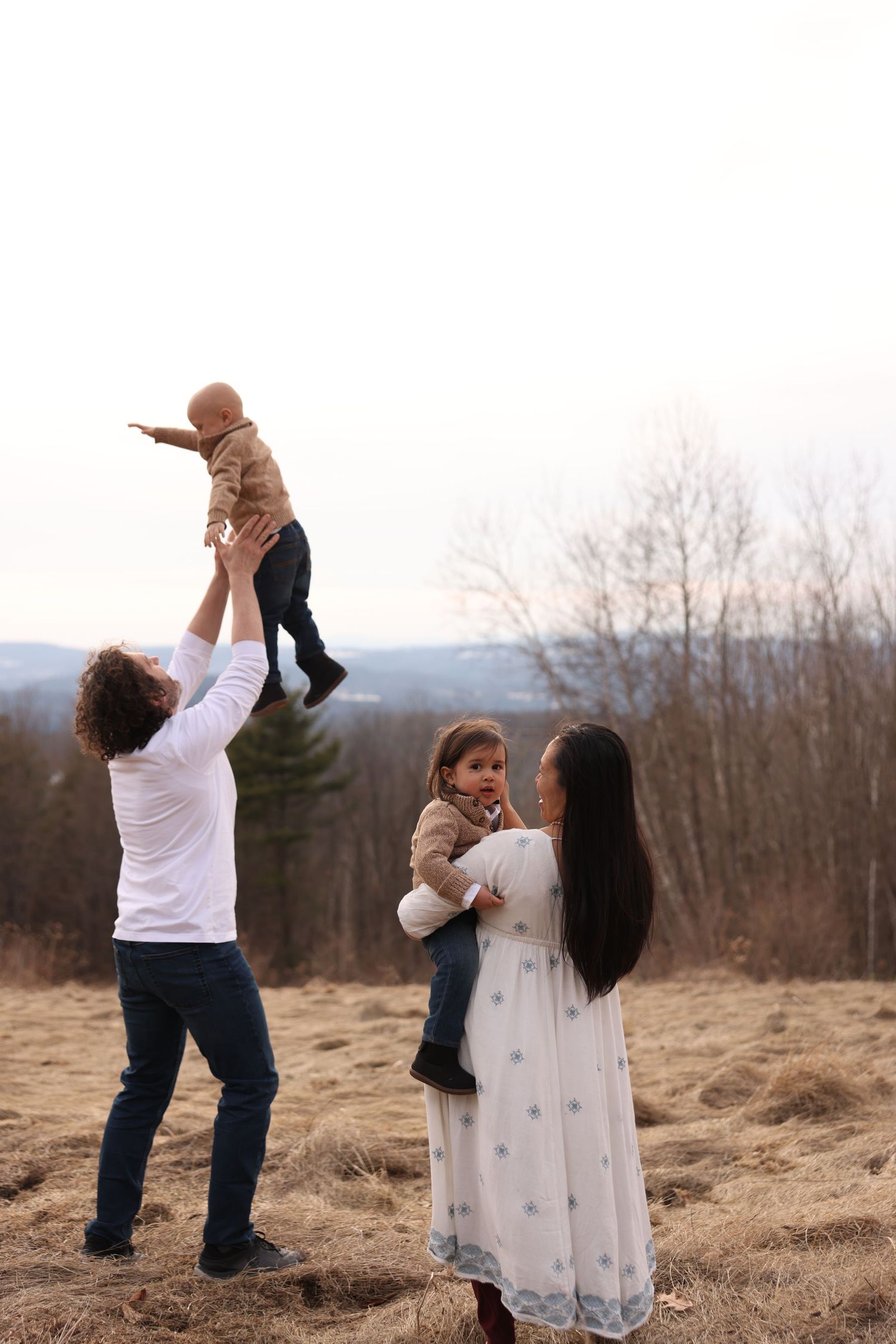 Father tossing baby in air while pregnant mother holds toddler in an open field outdoors.