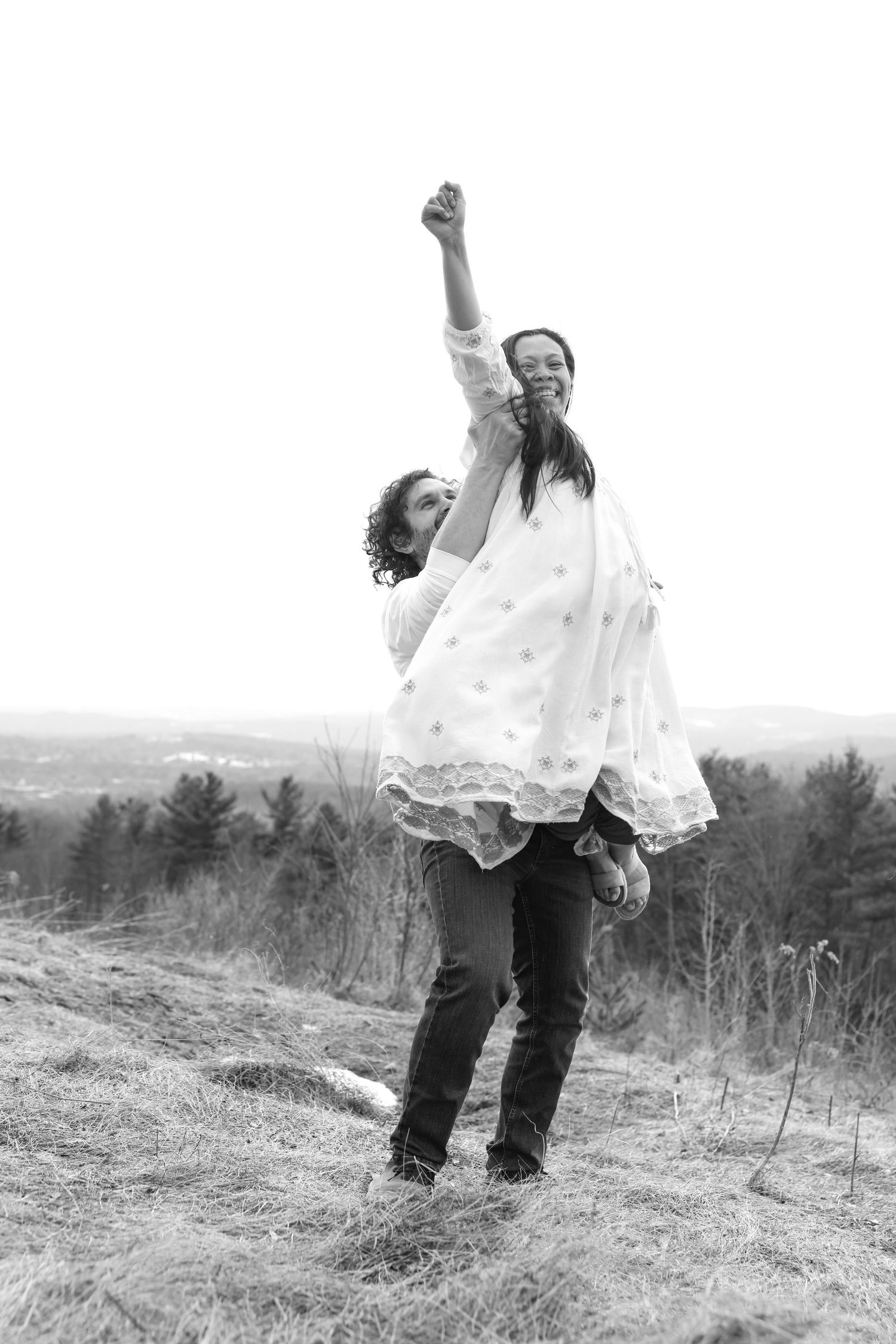 Black and white photo of man lifting smiling pregnant woman in white dress outdoors.