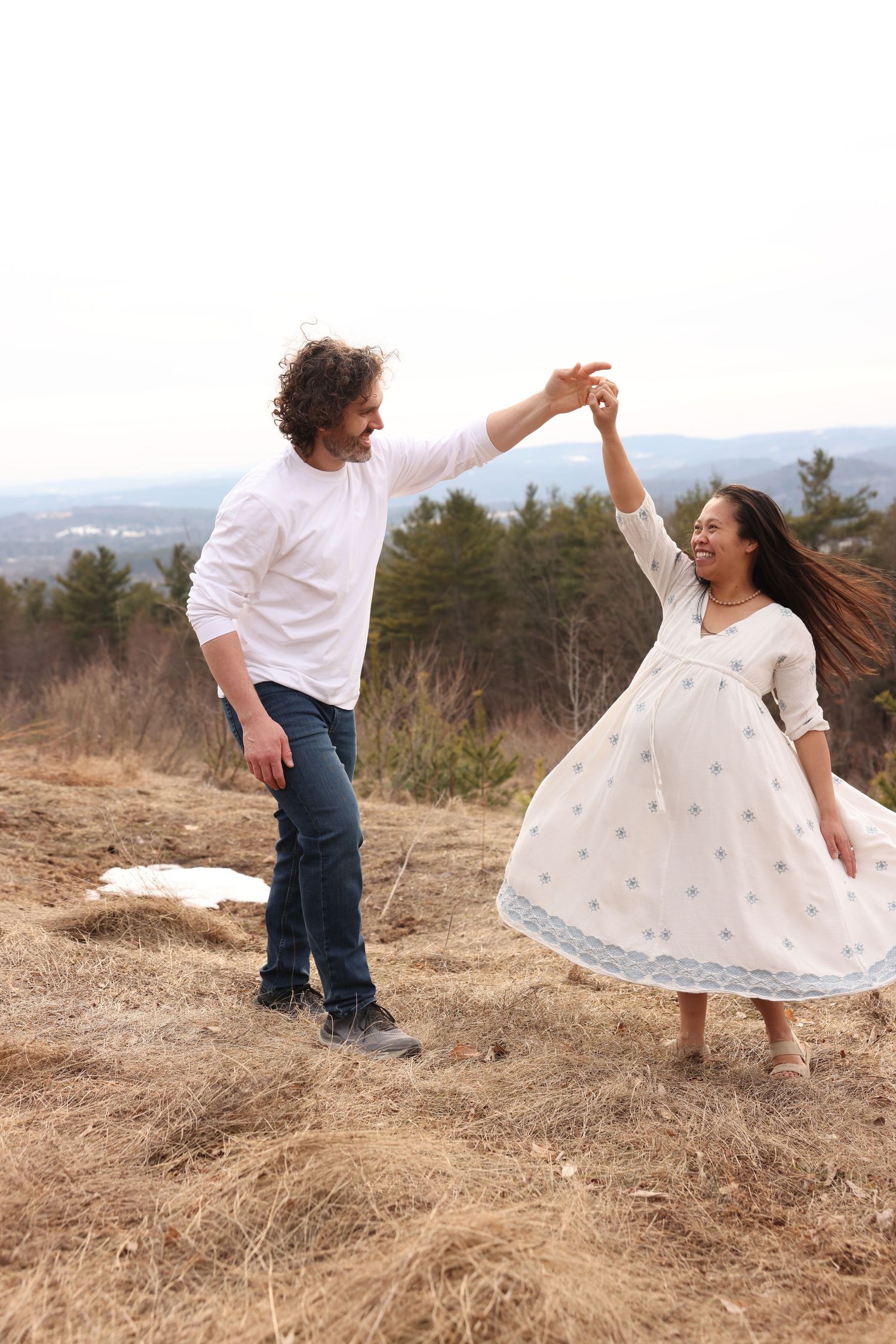 Couple dancing outdoors, man spinning pregnant woman in white dress on a hilltop meadow.