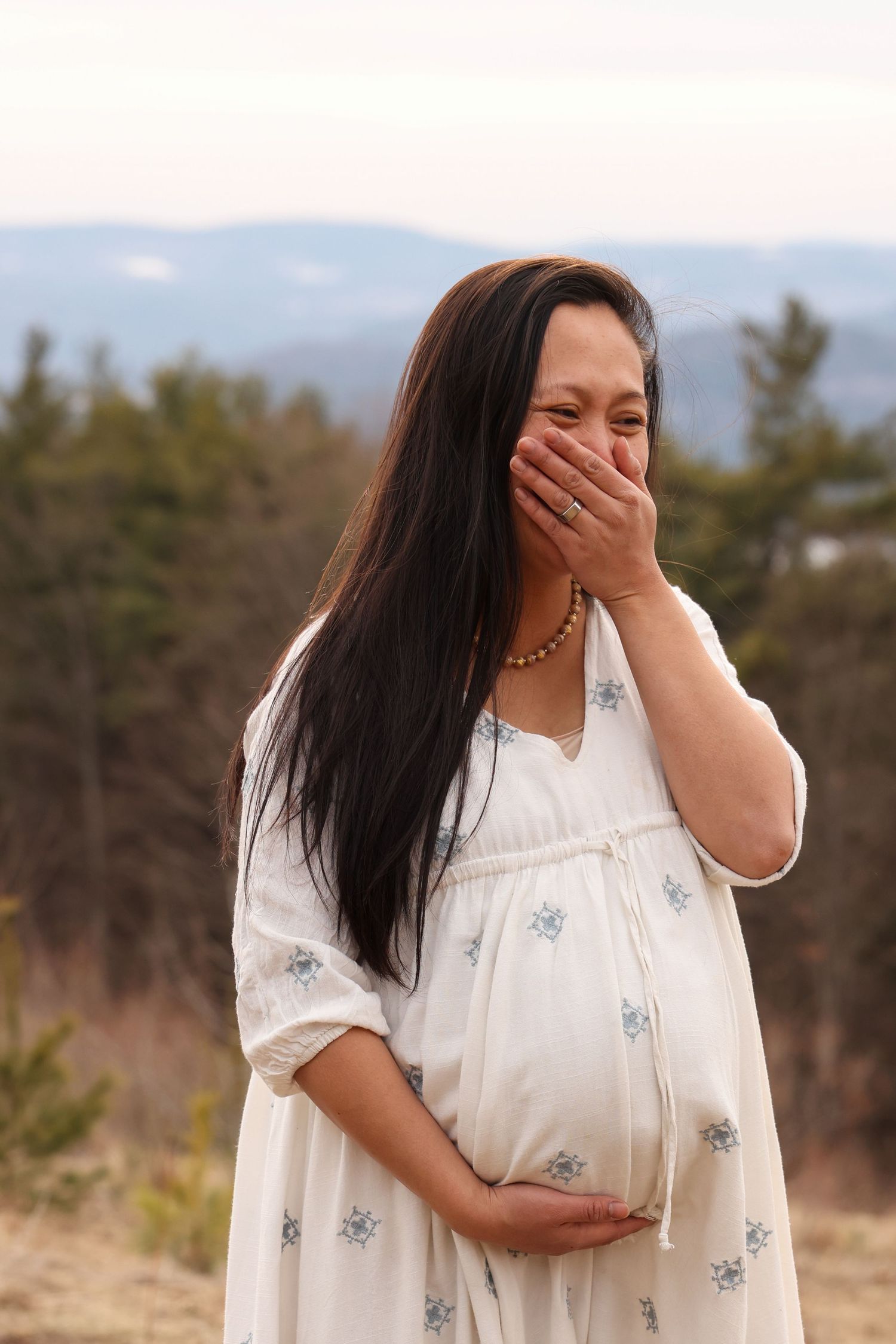 Pregnant woman in white floral dress laughing while holding belly on a scenic hilltop.