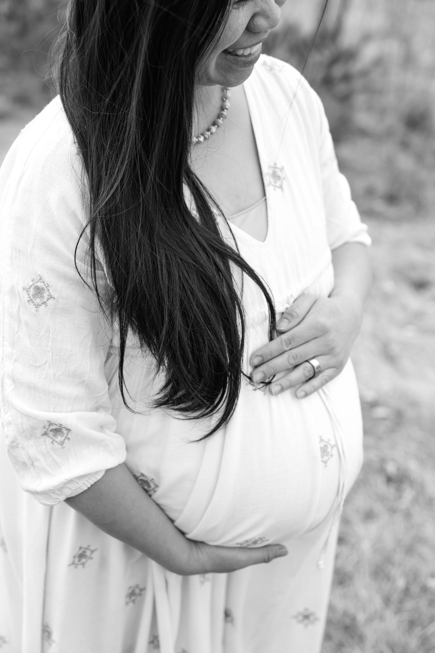Black and white close-up of smiling pregnant woman in white dress cradling her baby bump.