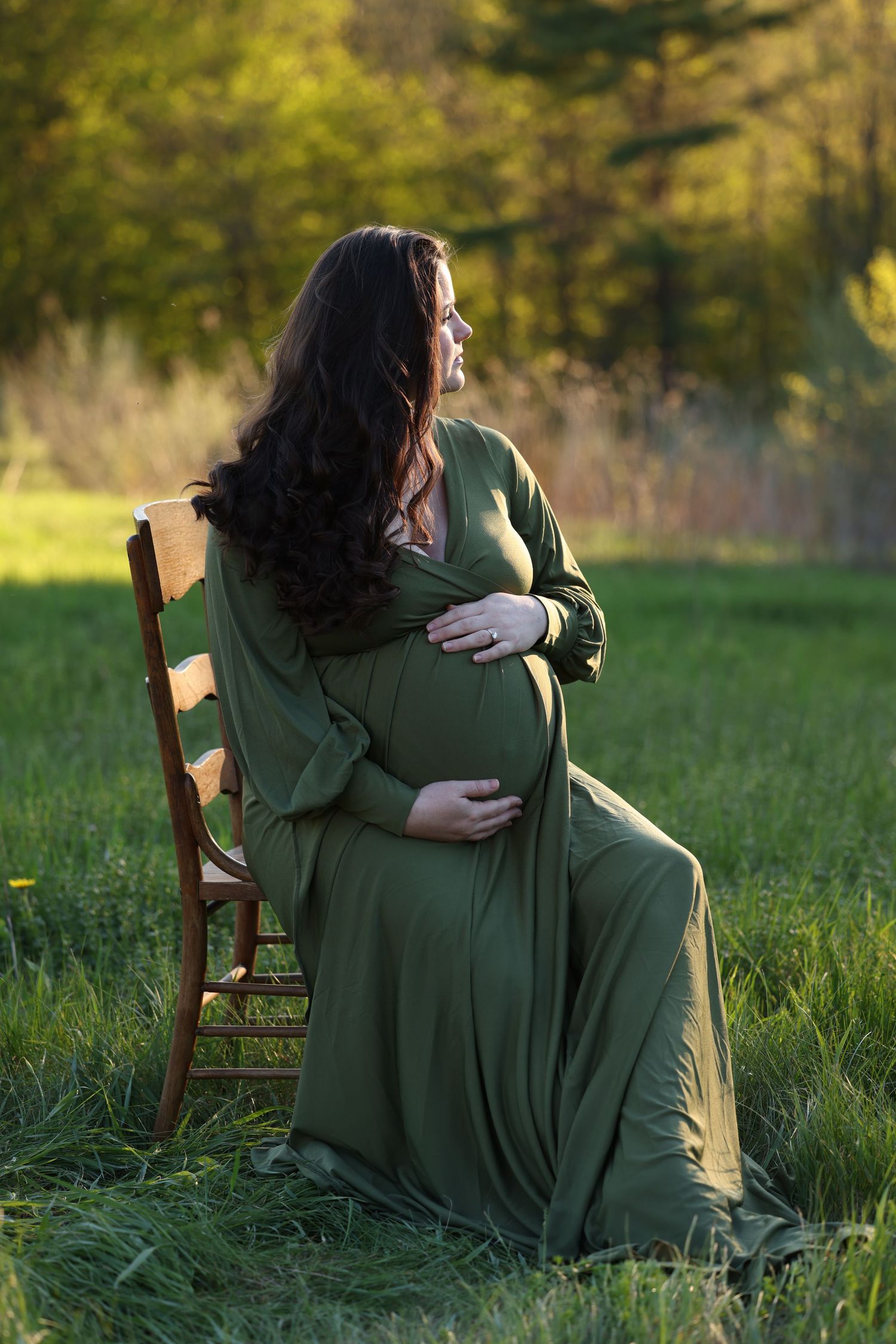 Pregnant woman in green dress sitting on chair outdoors at sunset.