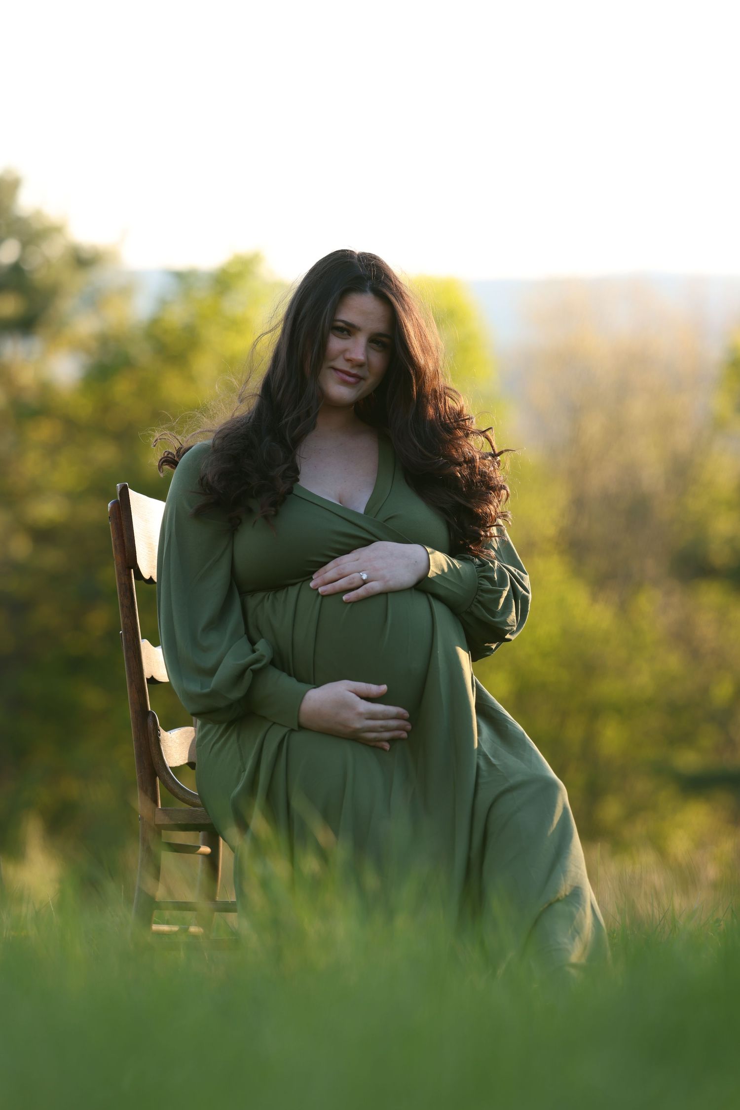 Pregnant woman in green dress sitting on chair outdoors, cradling baby bump in golden sunlight.