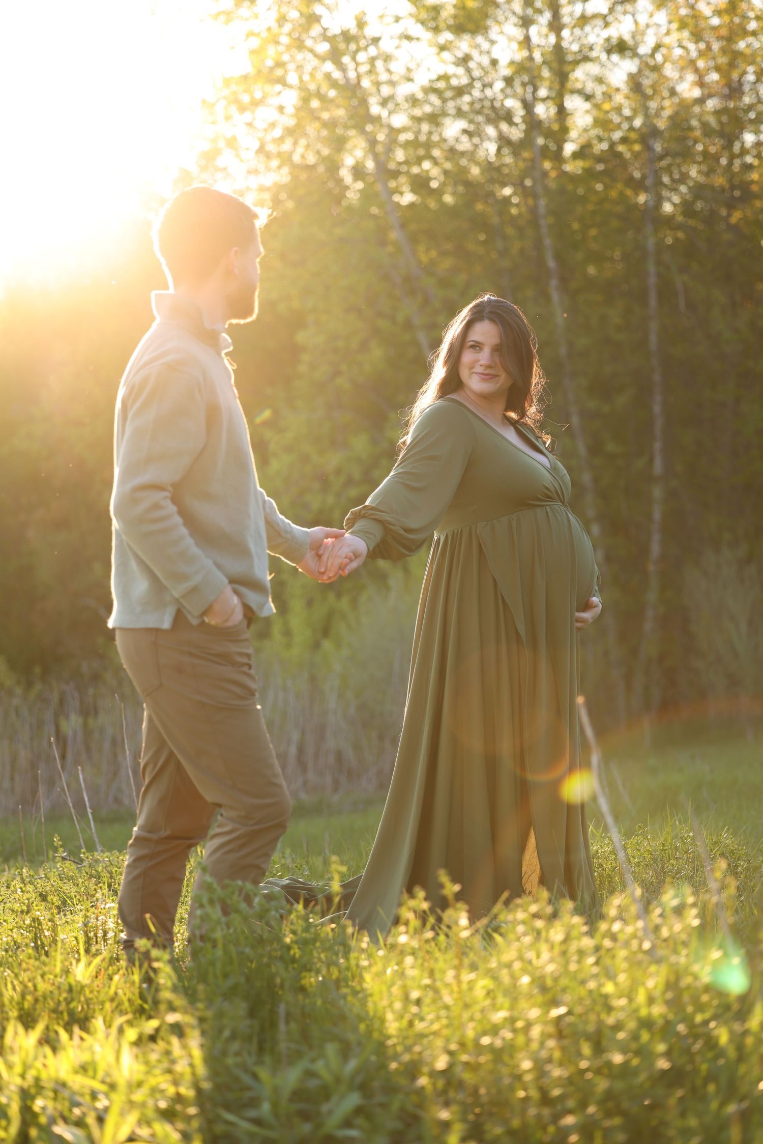 Pregnant woman in green dress holding hands with partner at sunset.