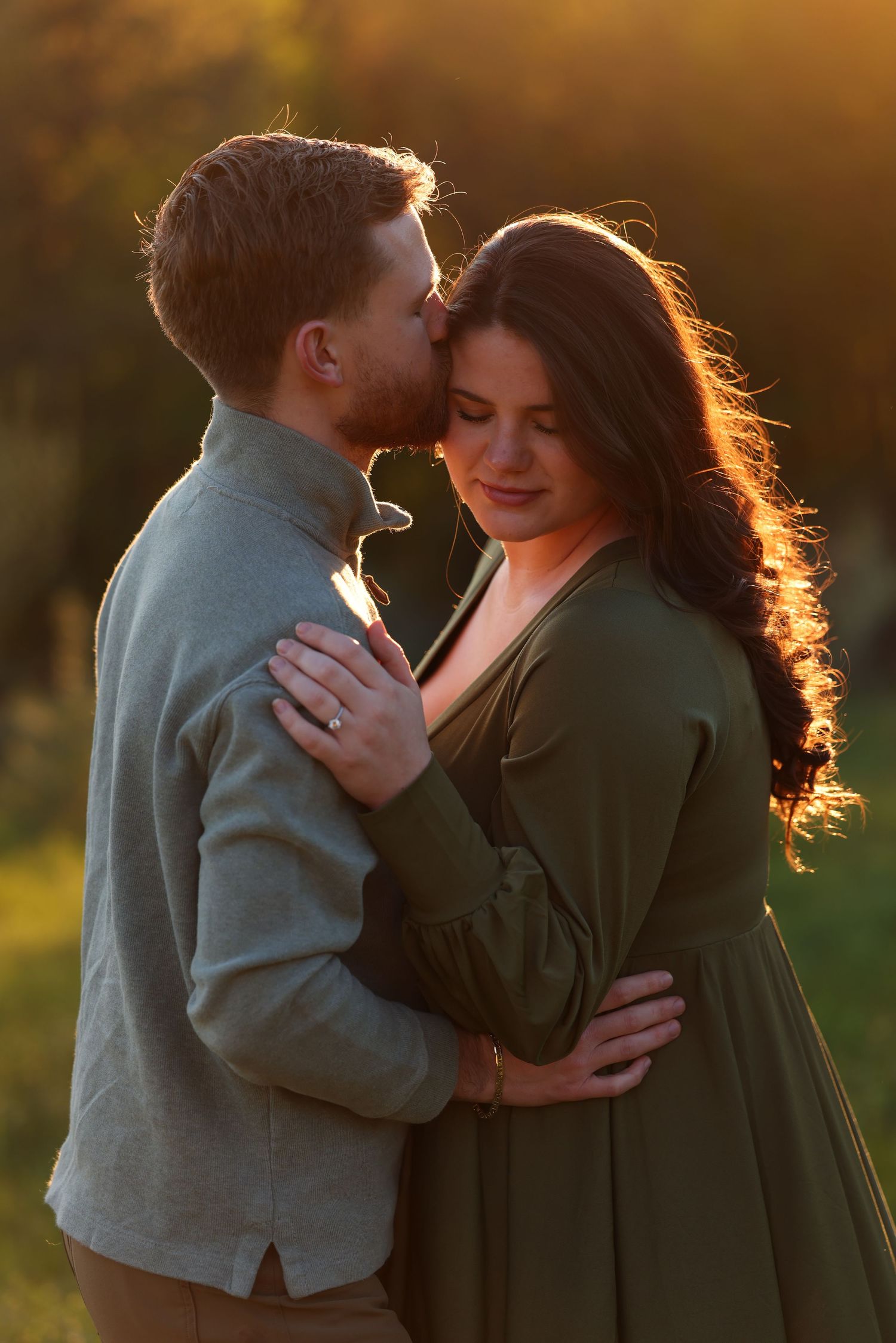 Man kissing pregnant woman's temple at golden hour outdoors.
