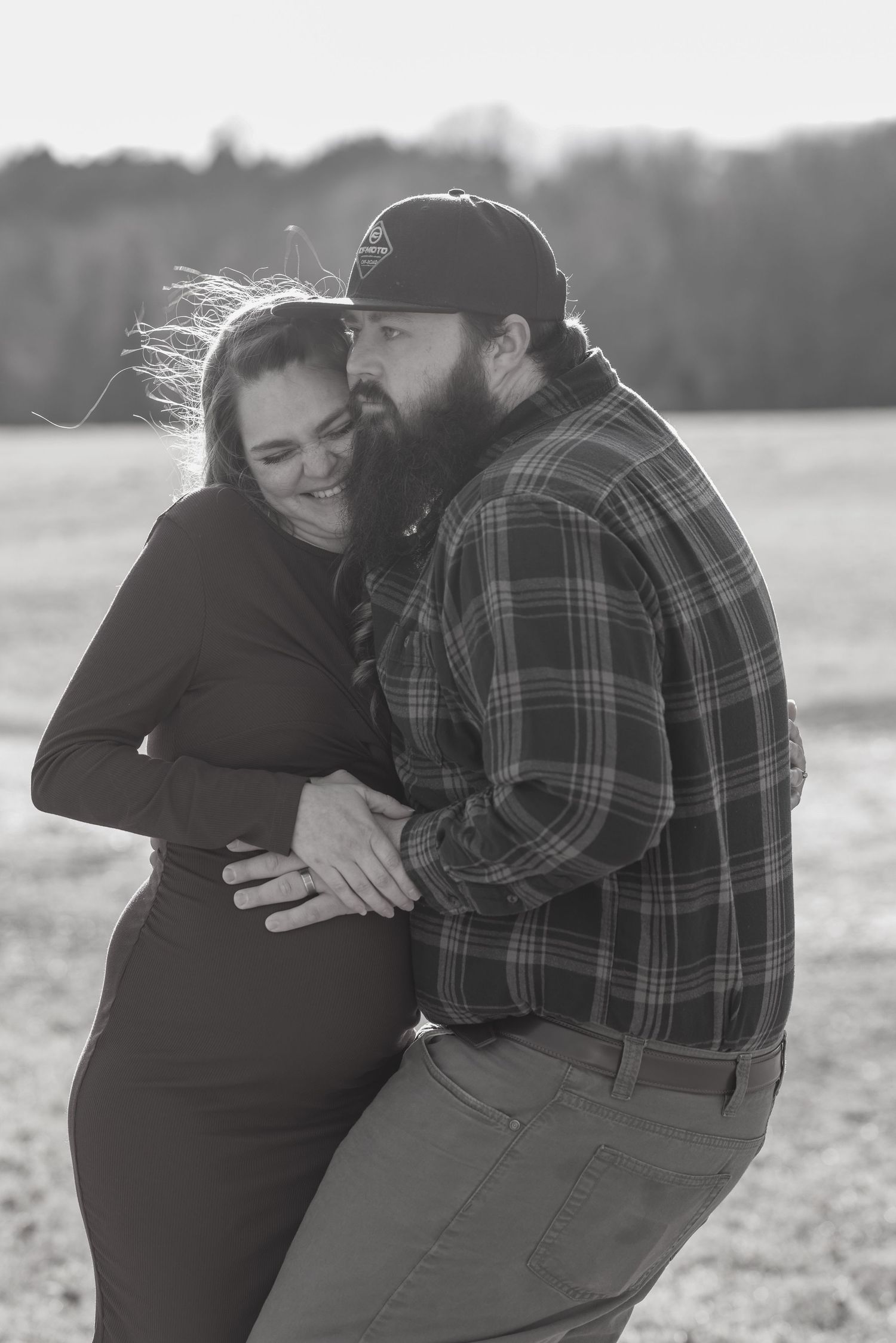 Black and white photo of bearded man hugging smiling pregnant woman in windy outdoor field.