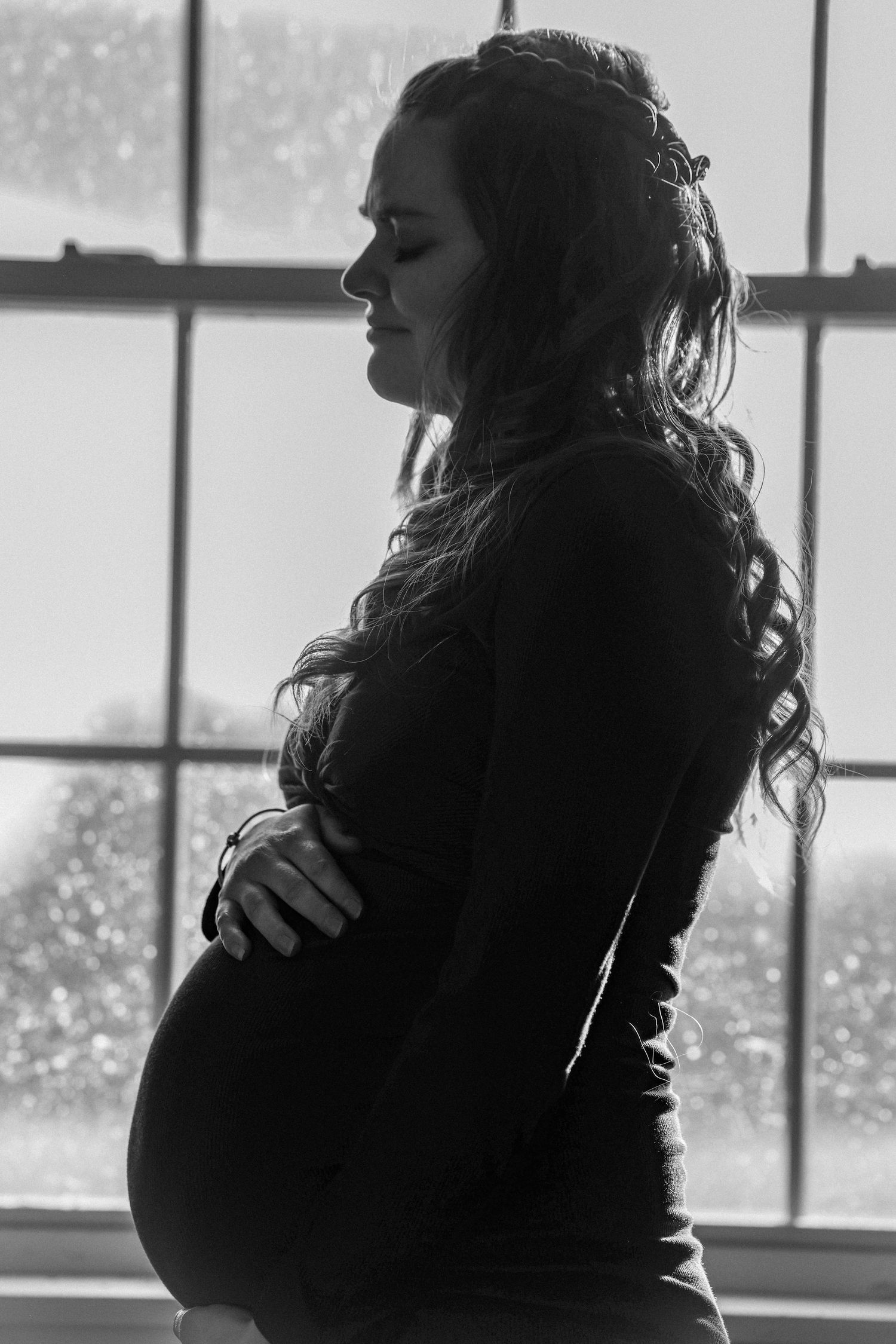 Black and white silhouette of pregnant woman cradling bump by large window with soft backlight.