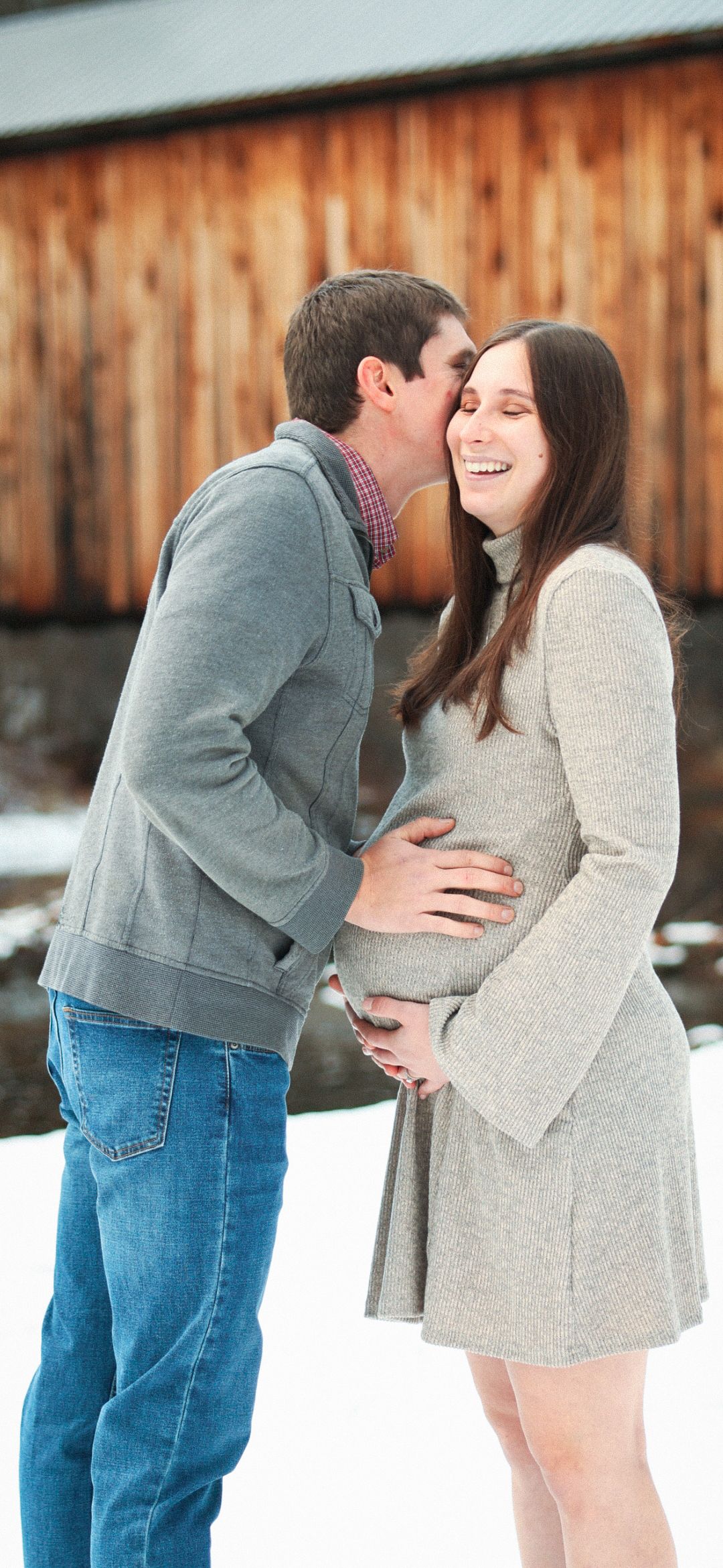 Couple laughing together outdoors in snow, man kissing pregnant woman's cheek near barn.