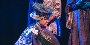 A performer in a glittering blue costume and ornate headpiece on stage during a theatrical production.